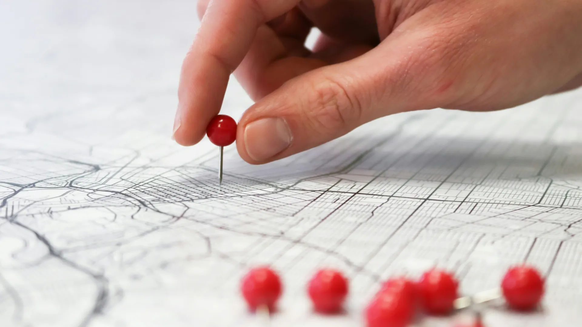 Hand placing red pushpin on a detailed map with other pins scattered nearby on a white surface.