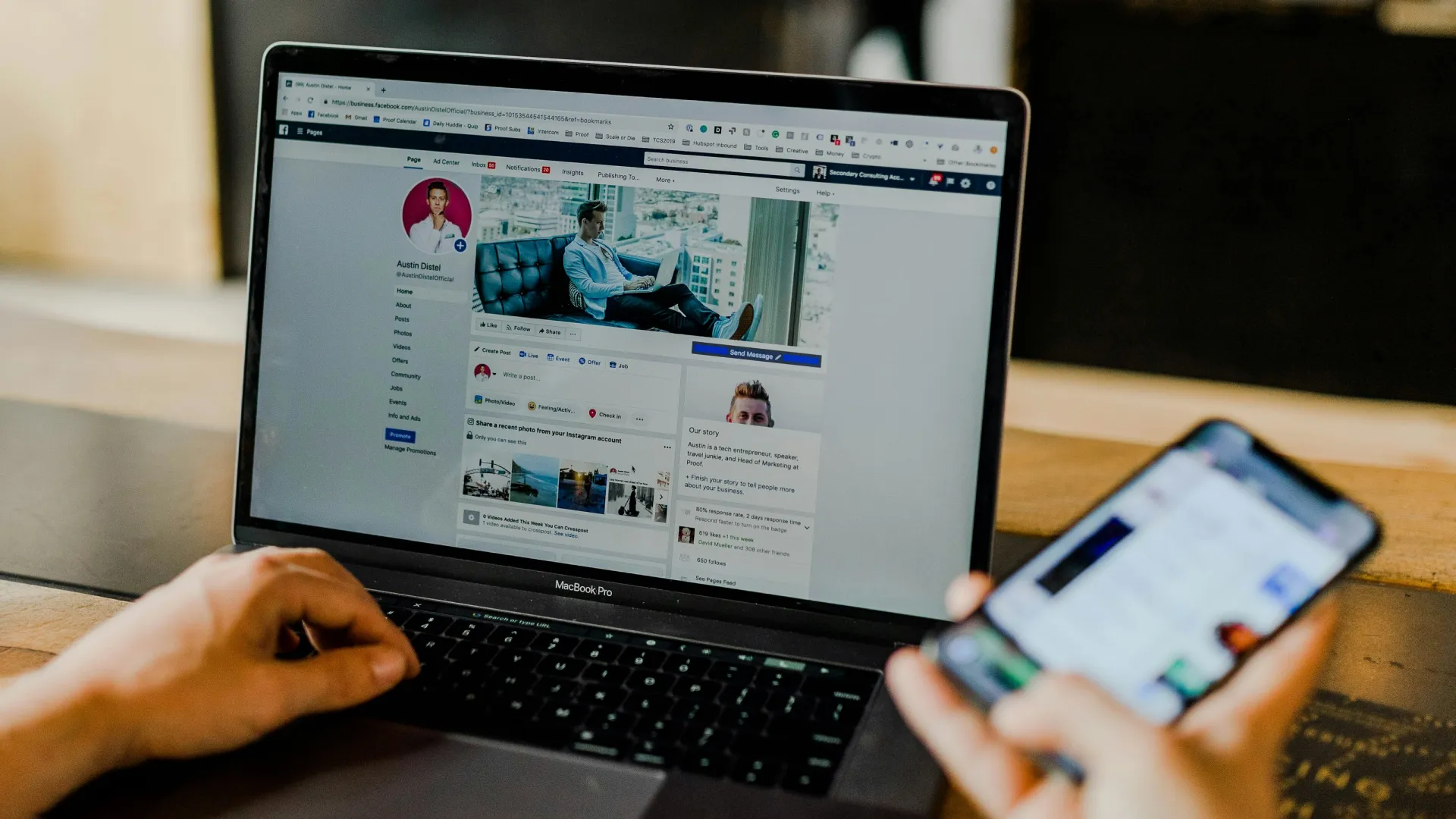 Person browsing a social media profile on a MacBook Pro laptop and smartphone at a wooden table.