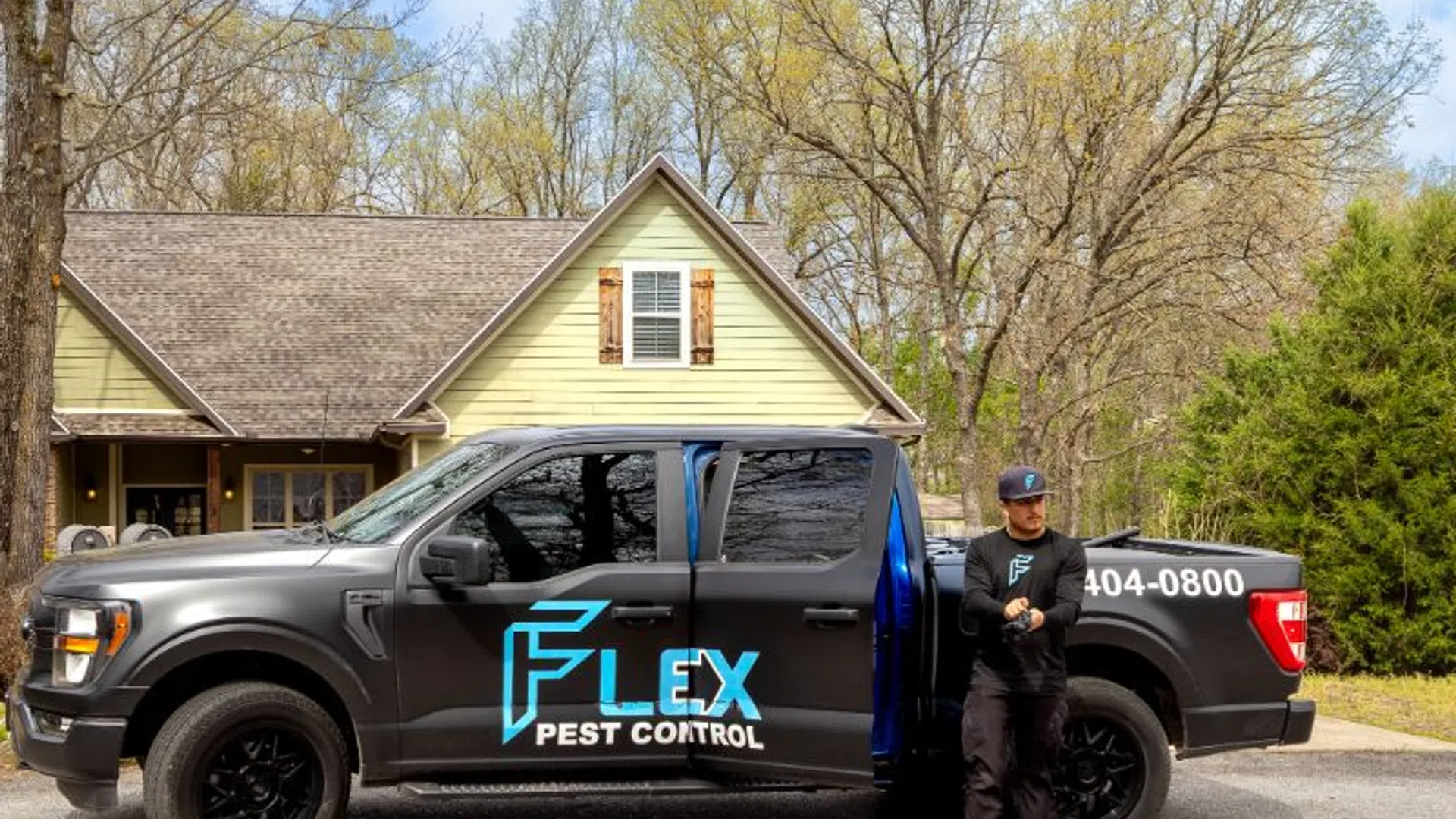 Black Flex Pest Control truck parked in front of a house with technician standing beside it in a wooded area.