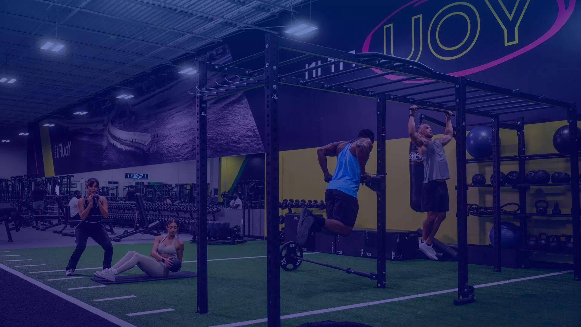 People exercising with weights and pull-up bars in a modern indoor gym with artificial turf and fitness equipment.