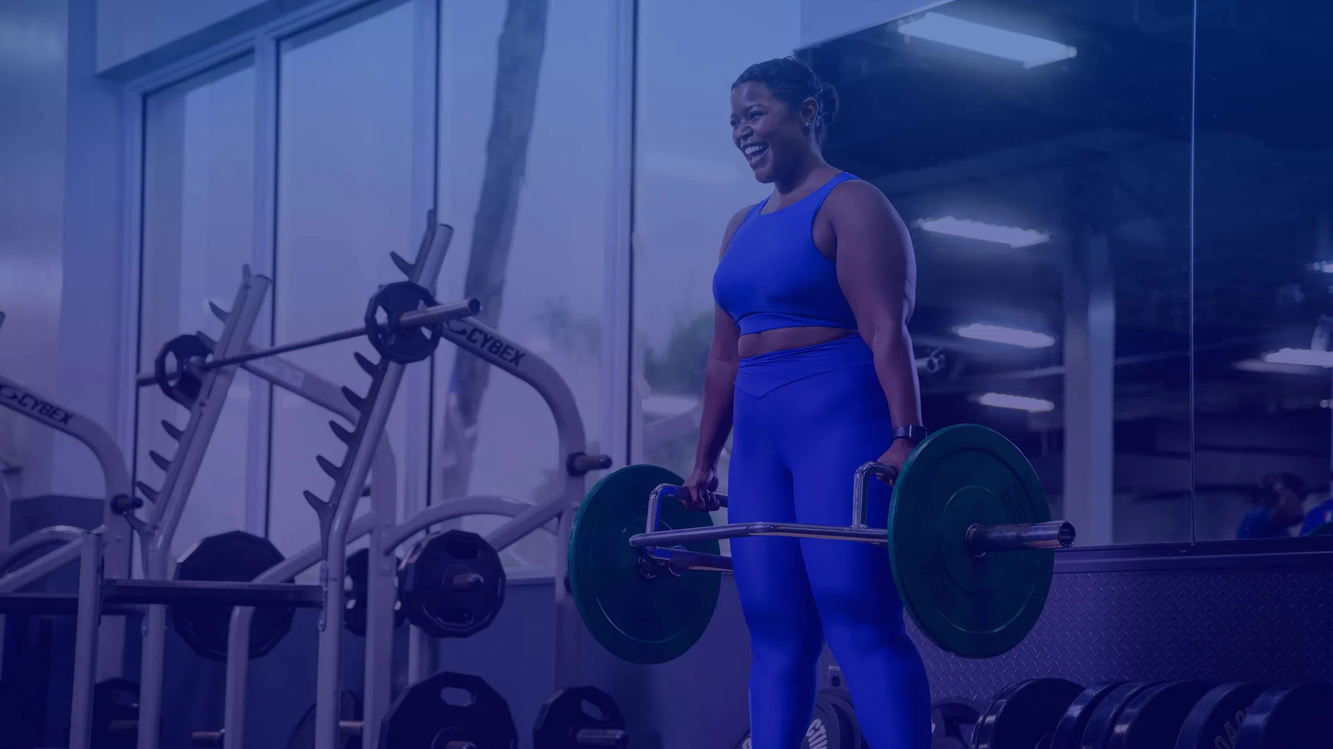 Woman in blue workout outfit lifting barbell with weights in gym setting with mirrors and equipment.