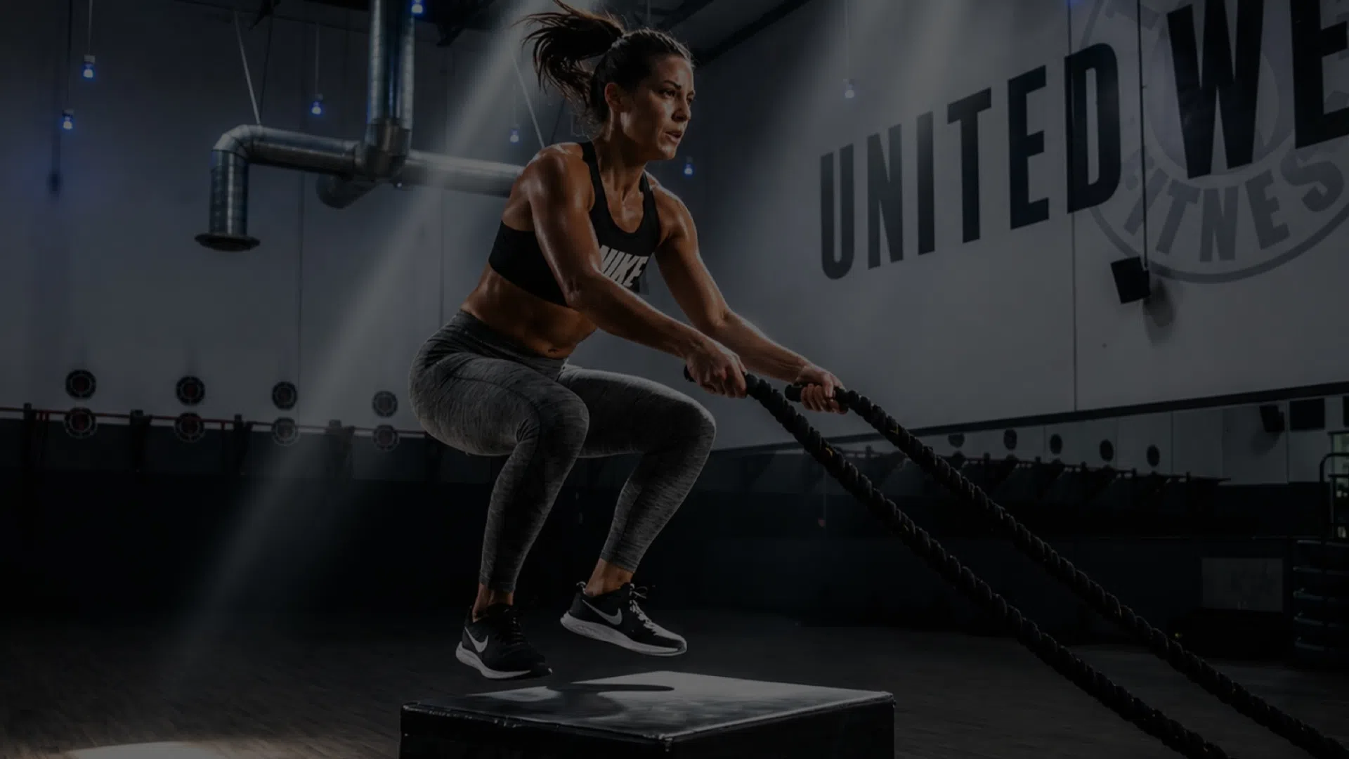 Athletic woman performing jump rope exercise on a box in a gym with industrial decor and United We Fitness sign.