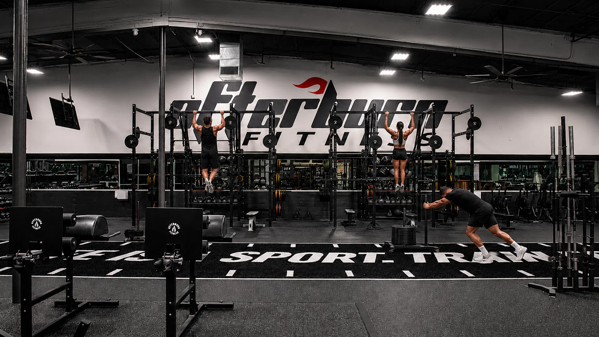 Three people exercising in a large gym with pull-up bars and fitness equipment under bright overhead lights.