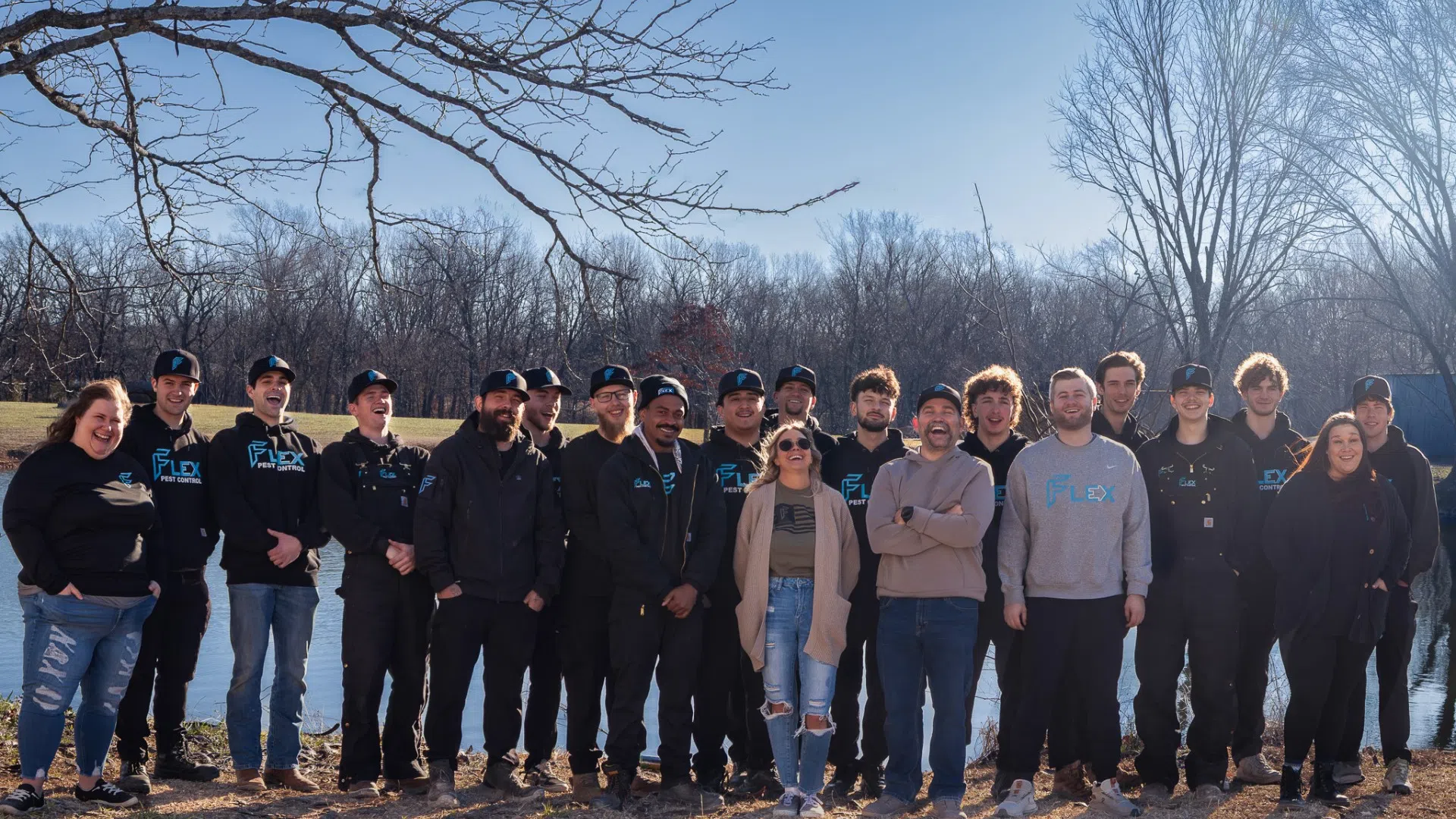 Group of casually dressed people standing outdoors by a lake on a clear sunny day with bare trees in the background