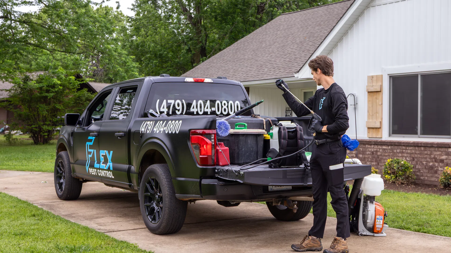 Pest control worker with equipment unloading from black Flex Pest Control truck in residential driveway.