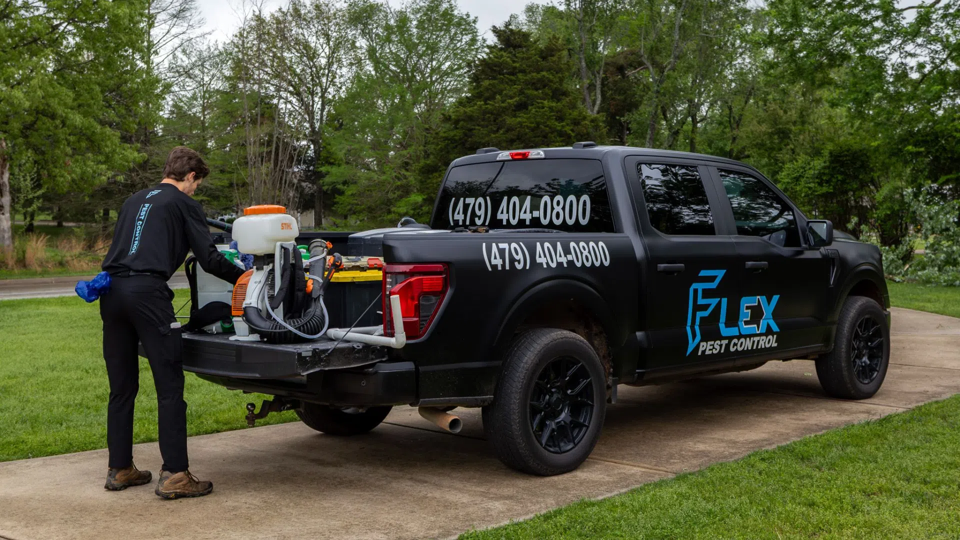 Pest control technician prepares equipment on black Flex Pest Control truck parked on driveway in green yard.