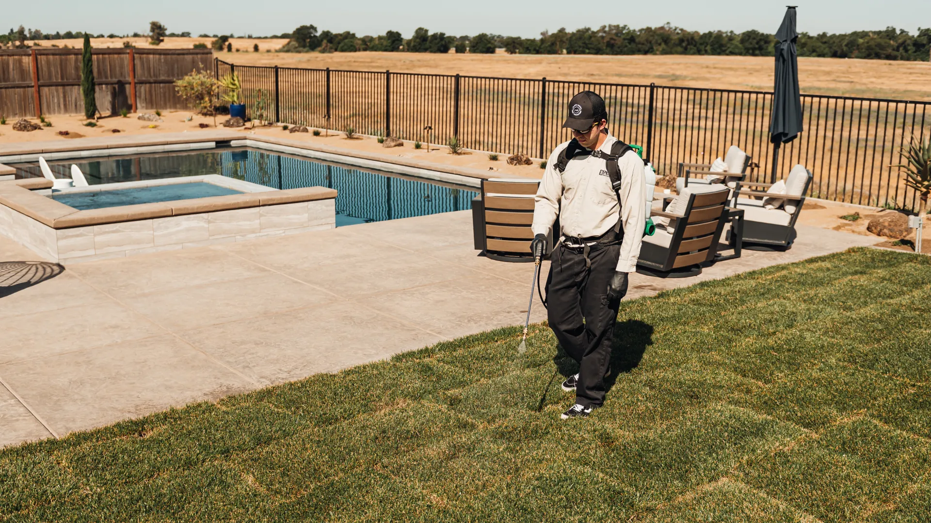 Technician wearing protective gear spraying pesticide on lawn near backyard pool and patio furniture in sunny yard.
