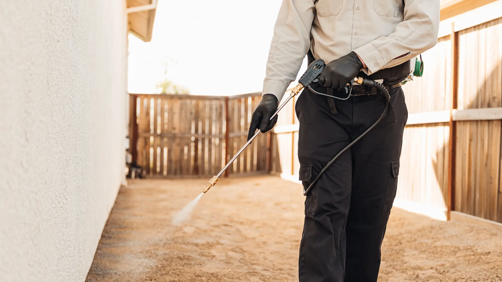 Pest control technician spraying insecticide outside a house near wooden fence on dirt ground.