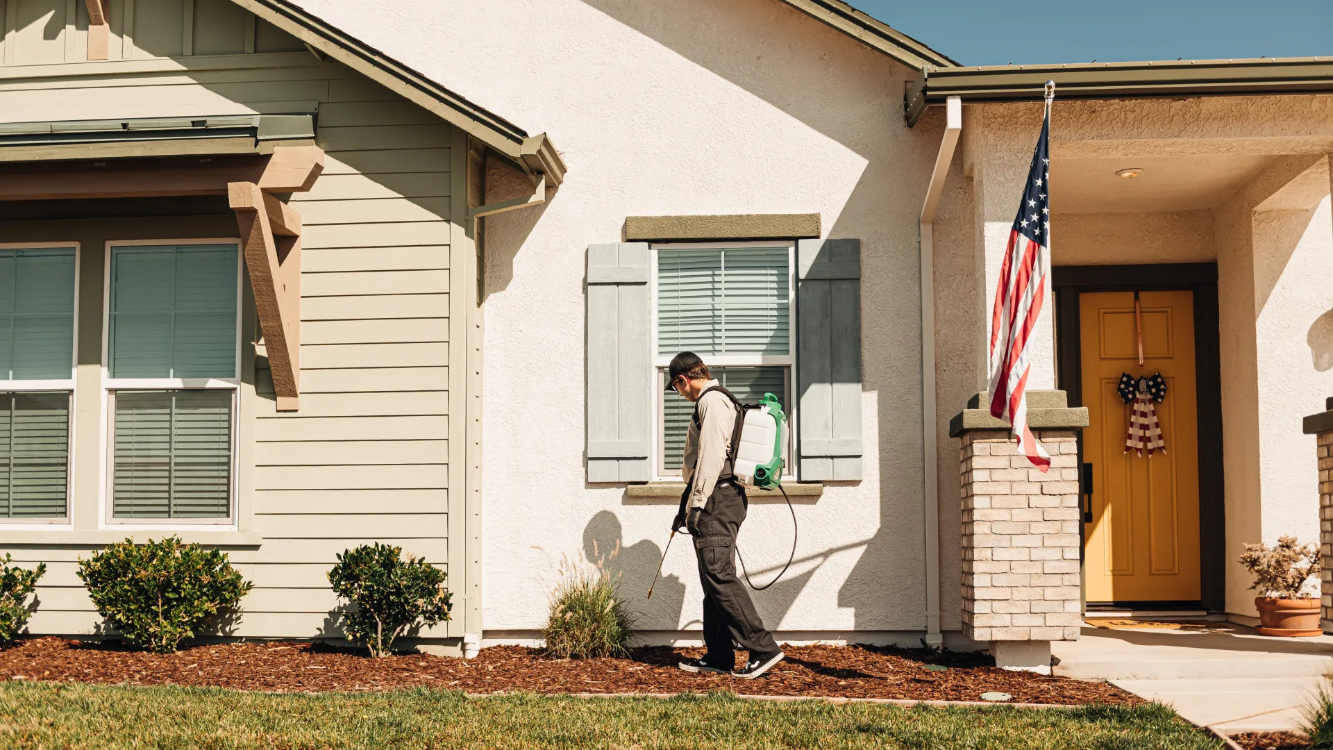 Pest control worker spraying insecticide around a suburban house with an American flag on the porch.