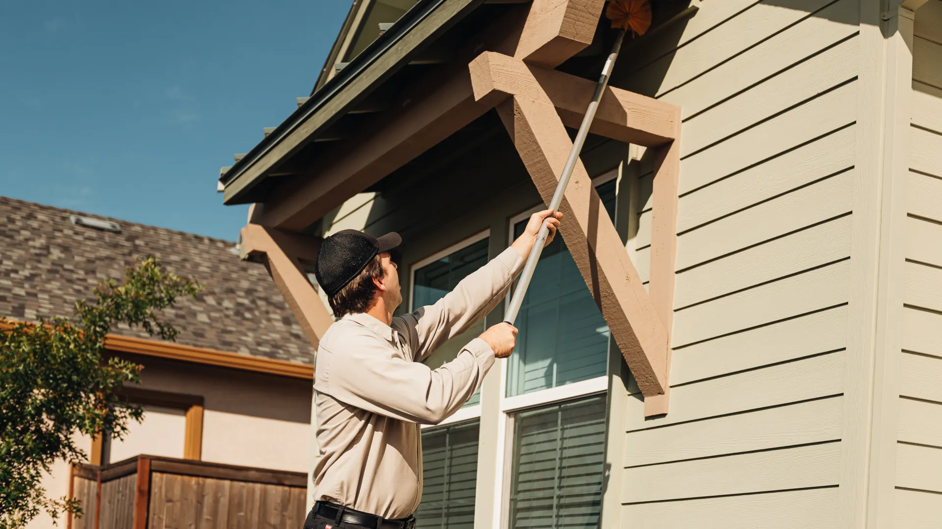 Man using a long-handled brush to clean exterior wooden trim on a house under clear sky.