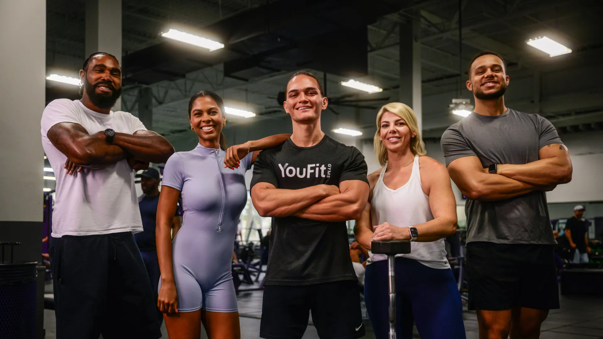 Five diverse fit adults confidently posing in gym workout attire with arms crossed and smiling in fitness center.