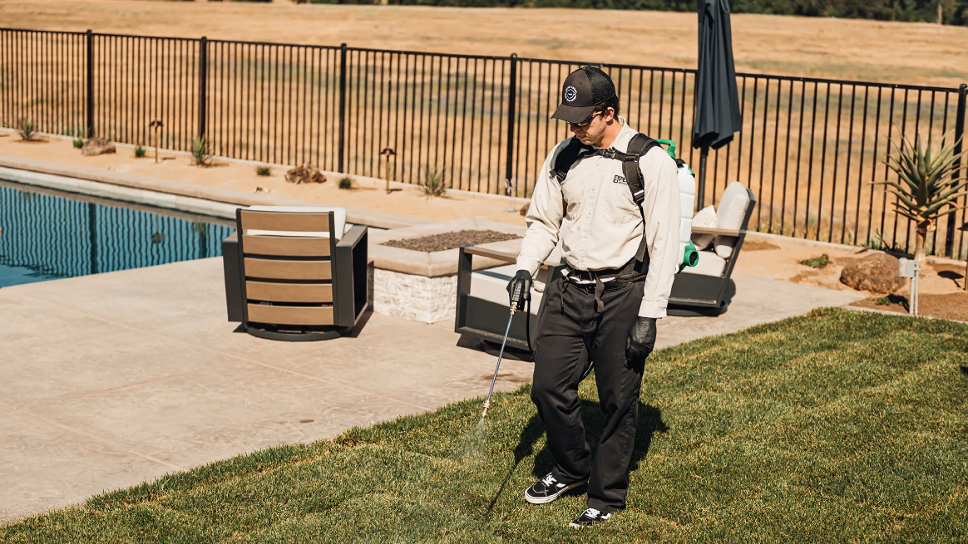Professional pest control technician spraying lawn near pool and patio area on sunny day.