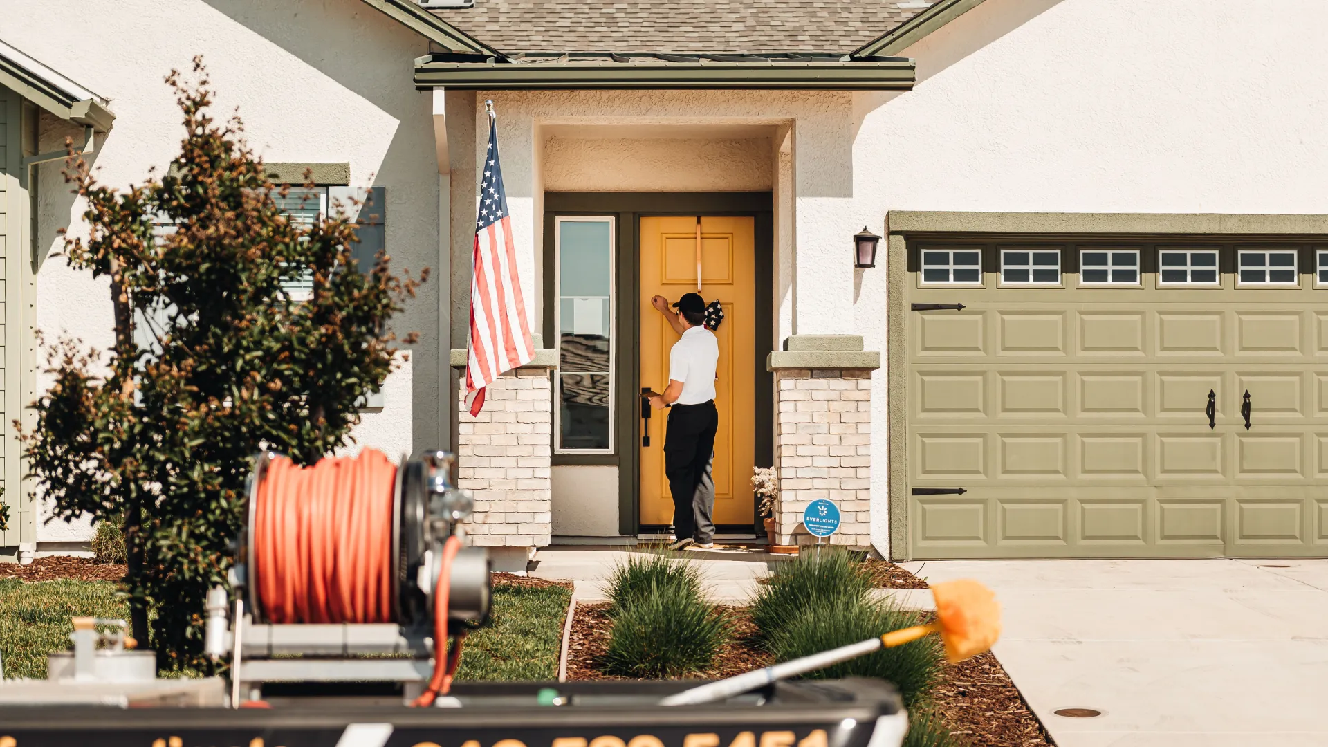 Technician knocking on front yellow door of suburban house beside an American flag.
