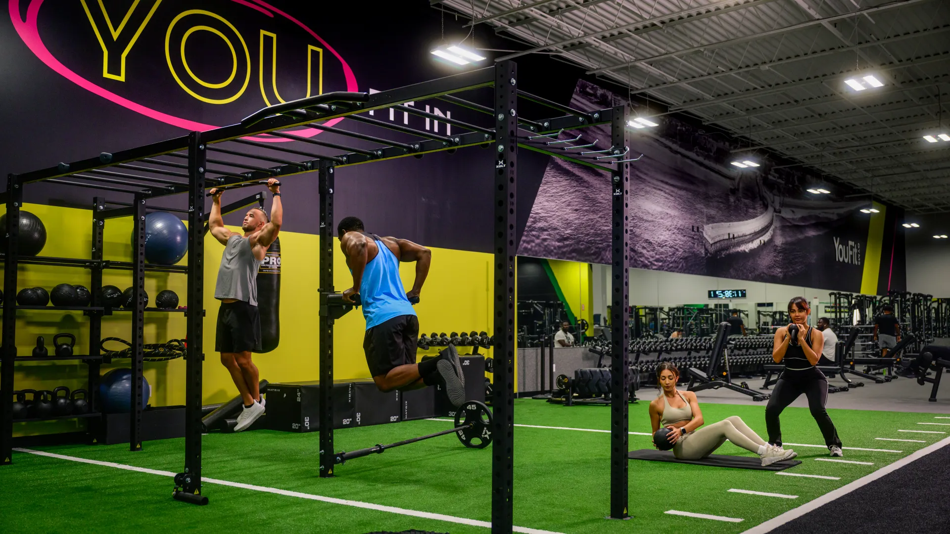 Two men performing pull-ups and ring dips on gym equipment while two women exercise on mat in a modern fitness center.