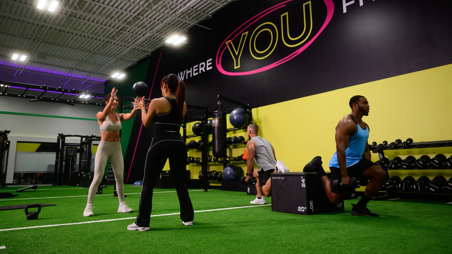 Four people exercising with medicine ball and weights in a modern gym with green turf and motivational wall text.