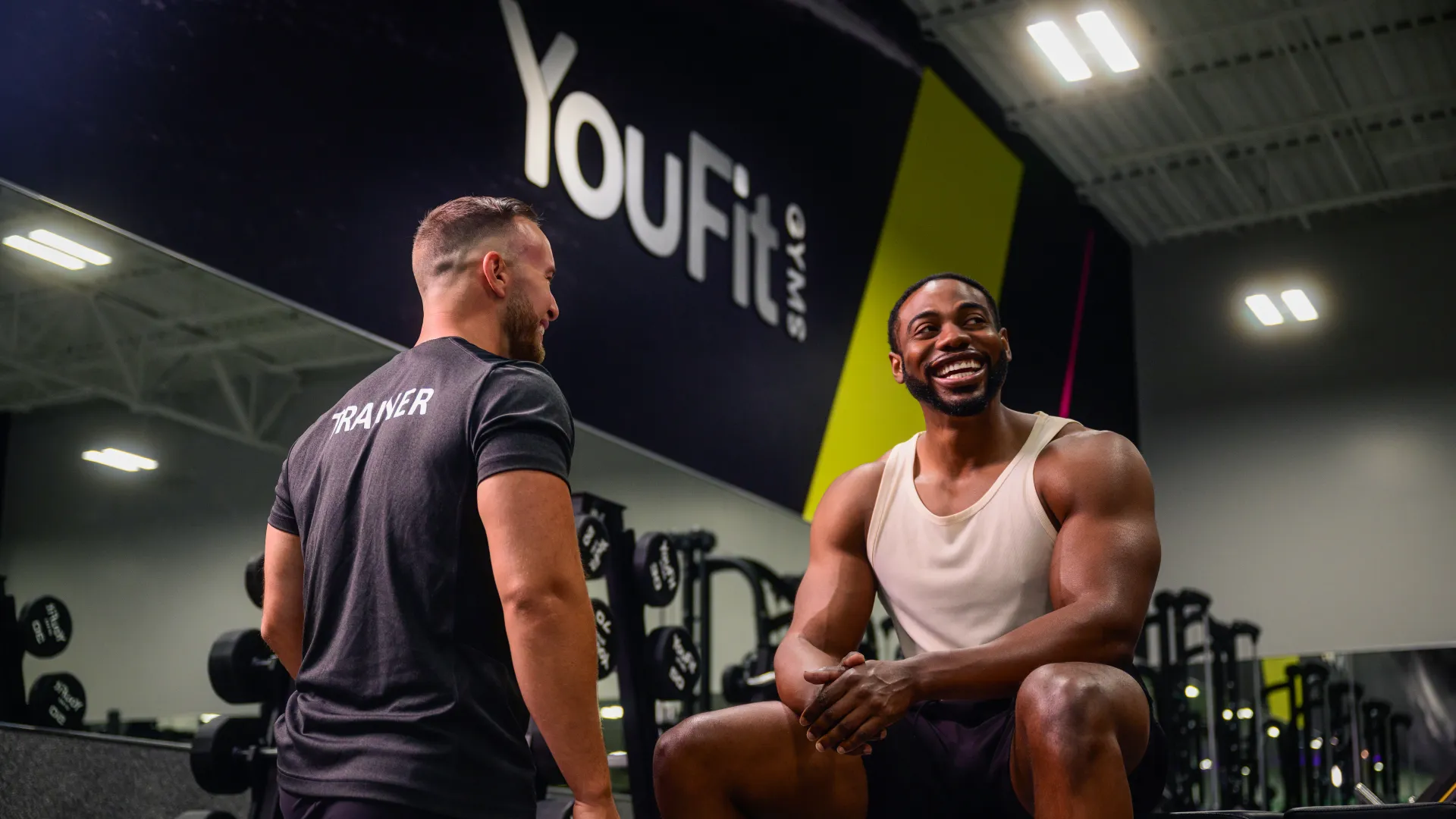 Smiling muscular man sitting on bench with trainer standing beside him at YouFit gym smiling and talking.