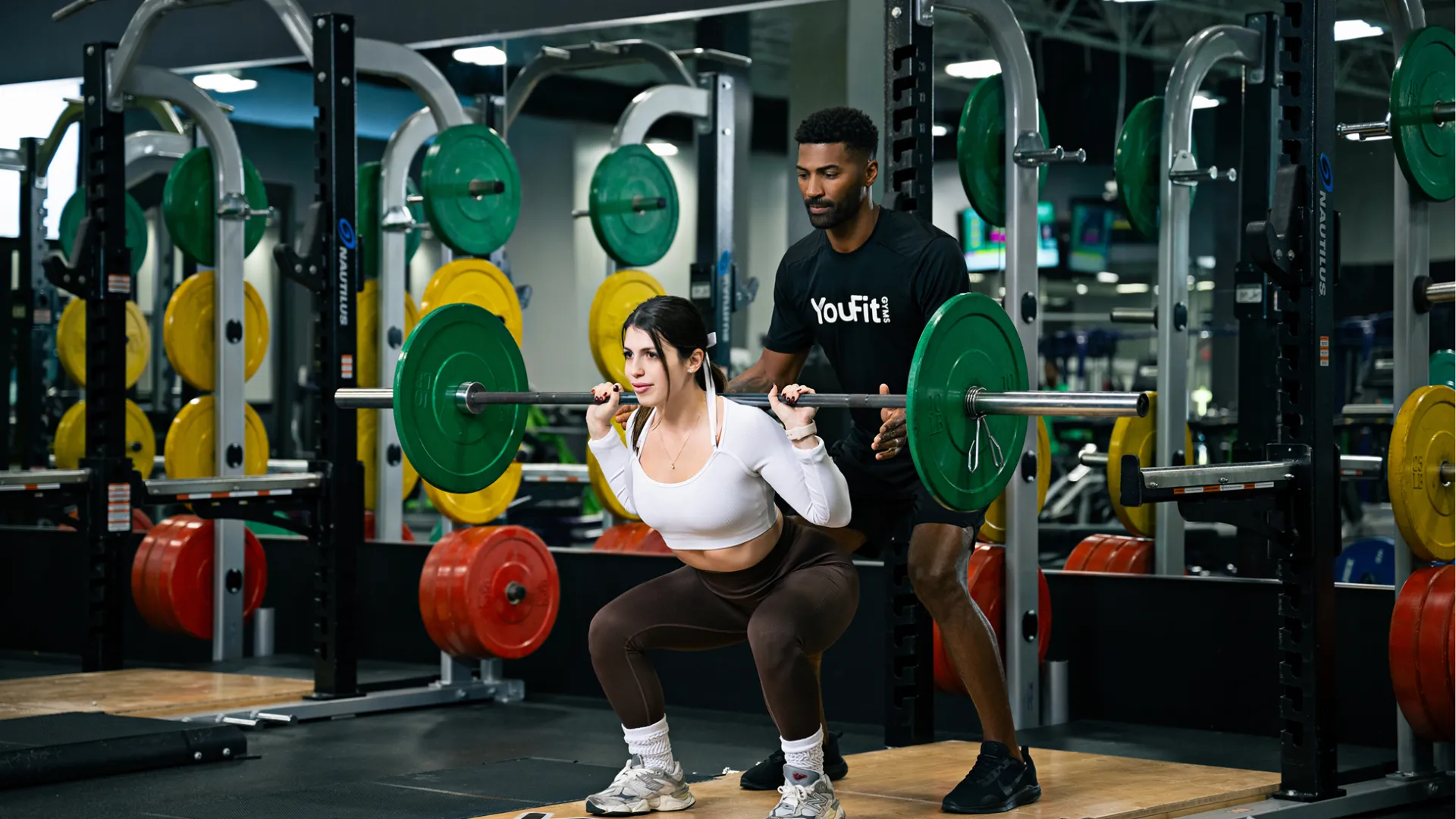 Female athlete performing barbell squat with male trainer spotting her in a gym with colorful weight plates.