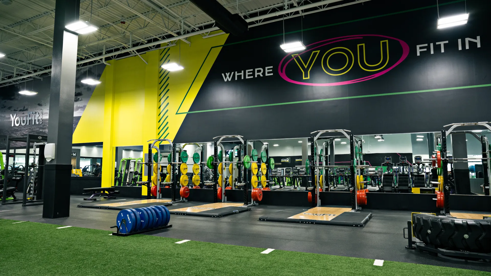 Modern gym interior with weightlifting racks, colorful weight plates, green turf, and motivational wall text.