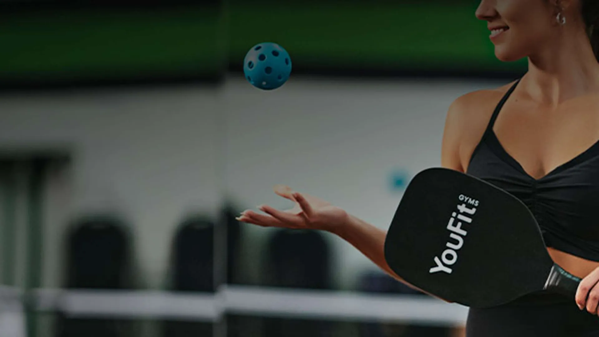 Woman in black workout attire playing pickleball with YouFit paddle and blue ball in indoor gym