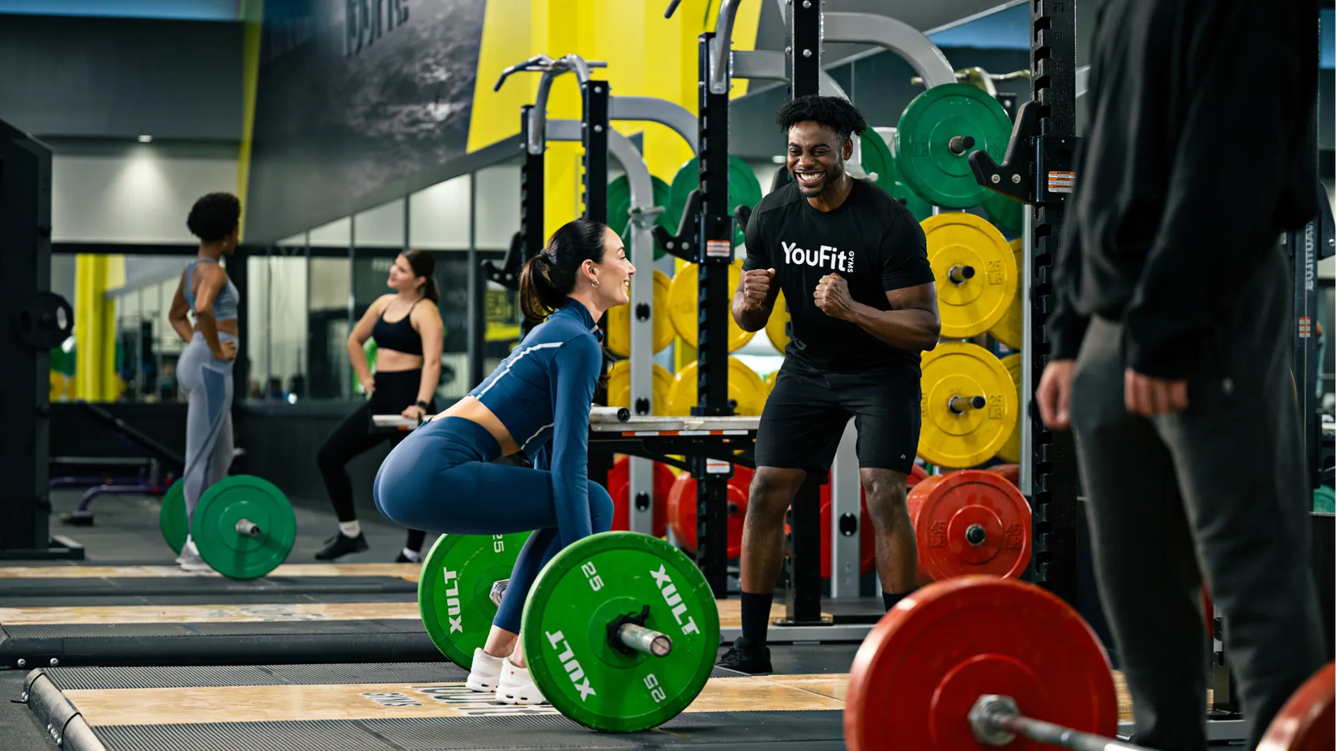 Woman lifting barbell with green weights coached by trainer in busy gym with colorful plates and mirrors