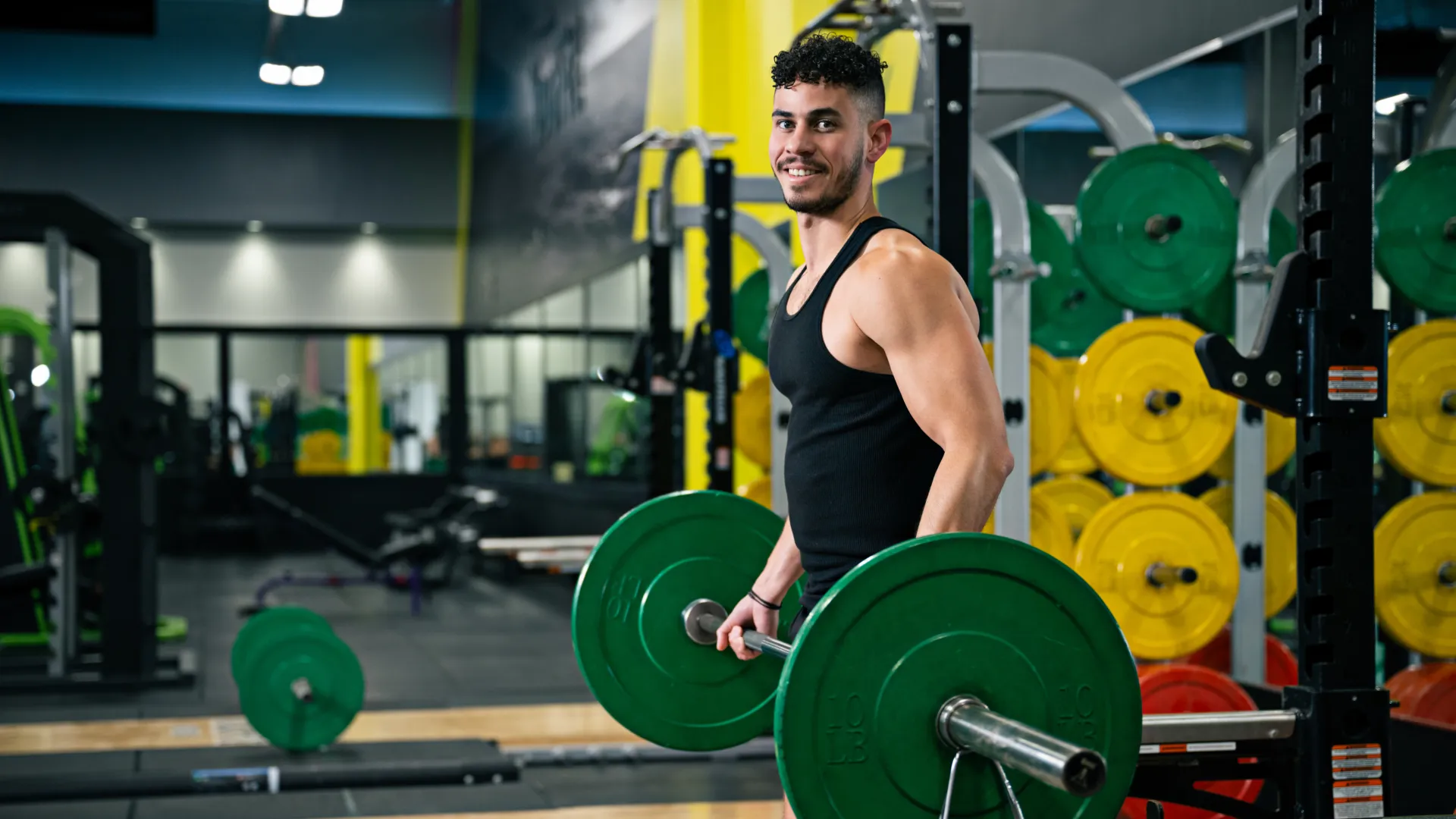 Smiling man in black tank top lifting heavy barbell weights in a gym with colorful weight plates in background