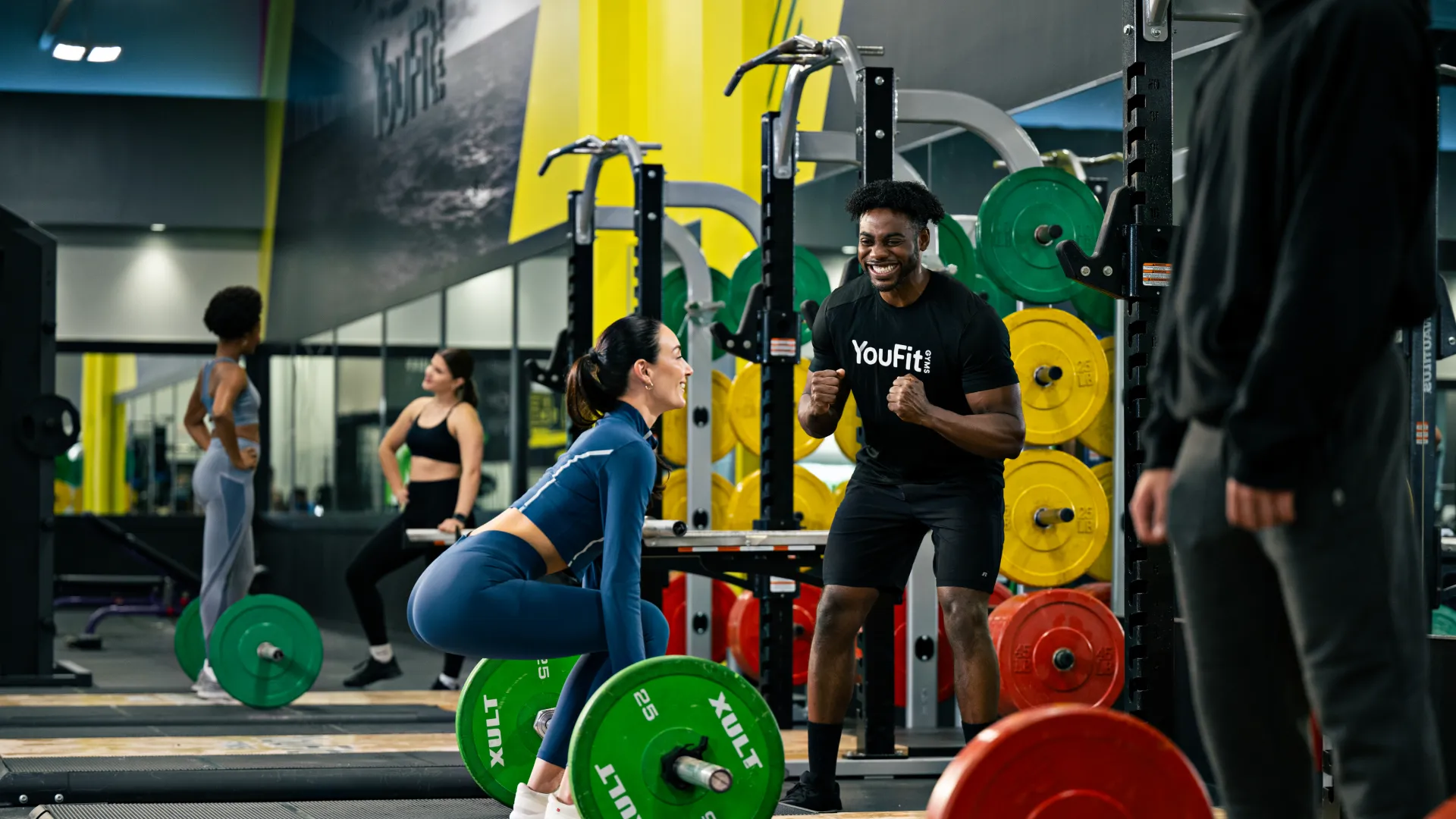 Woman lifting green barbell weights during training session in gym with coach motivating her.