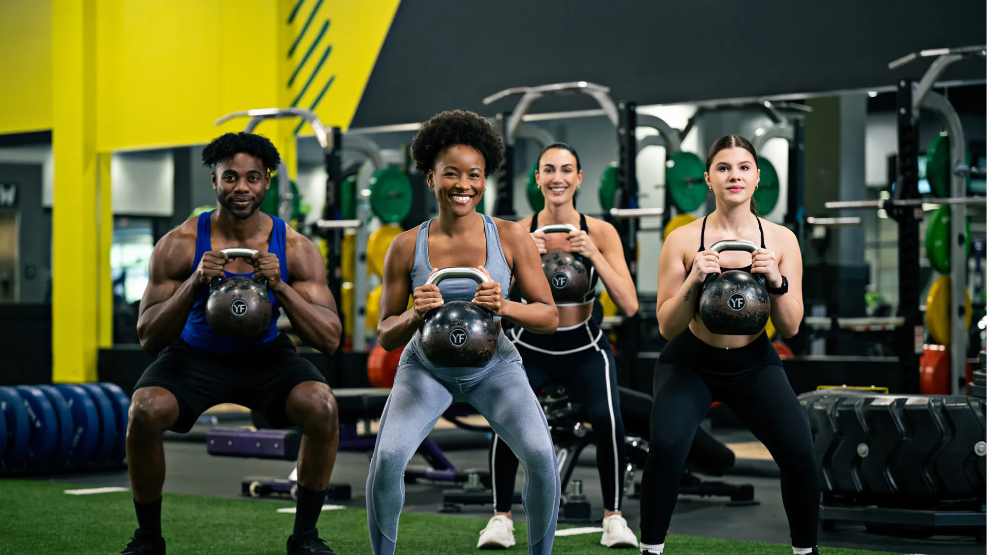 Group of four diverse adults performing kettlebell squats together in a modern gym setting.