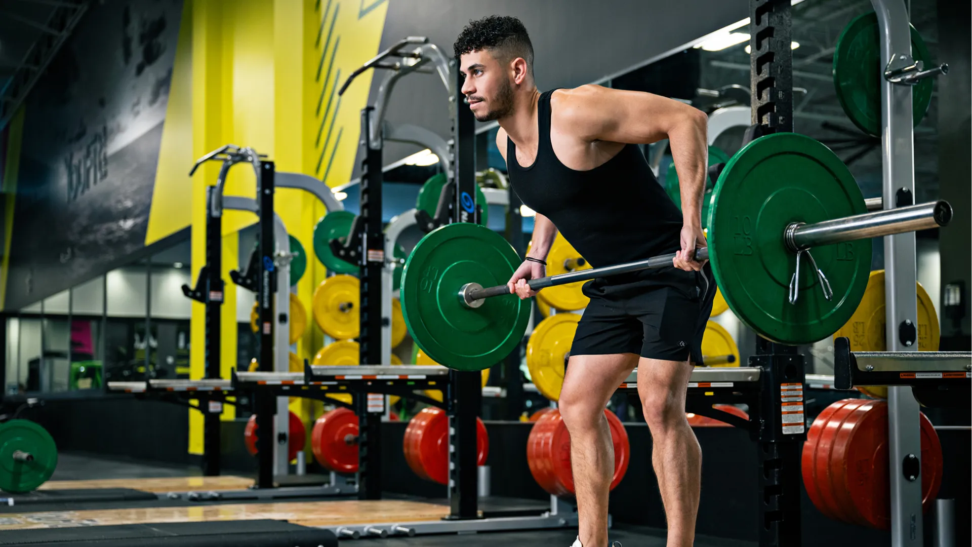 Man lifting barbell with green plates performing bent-over row exercise in modern gym with colorful weights.