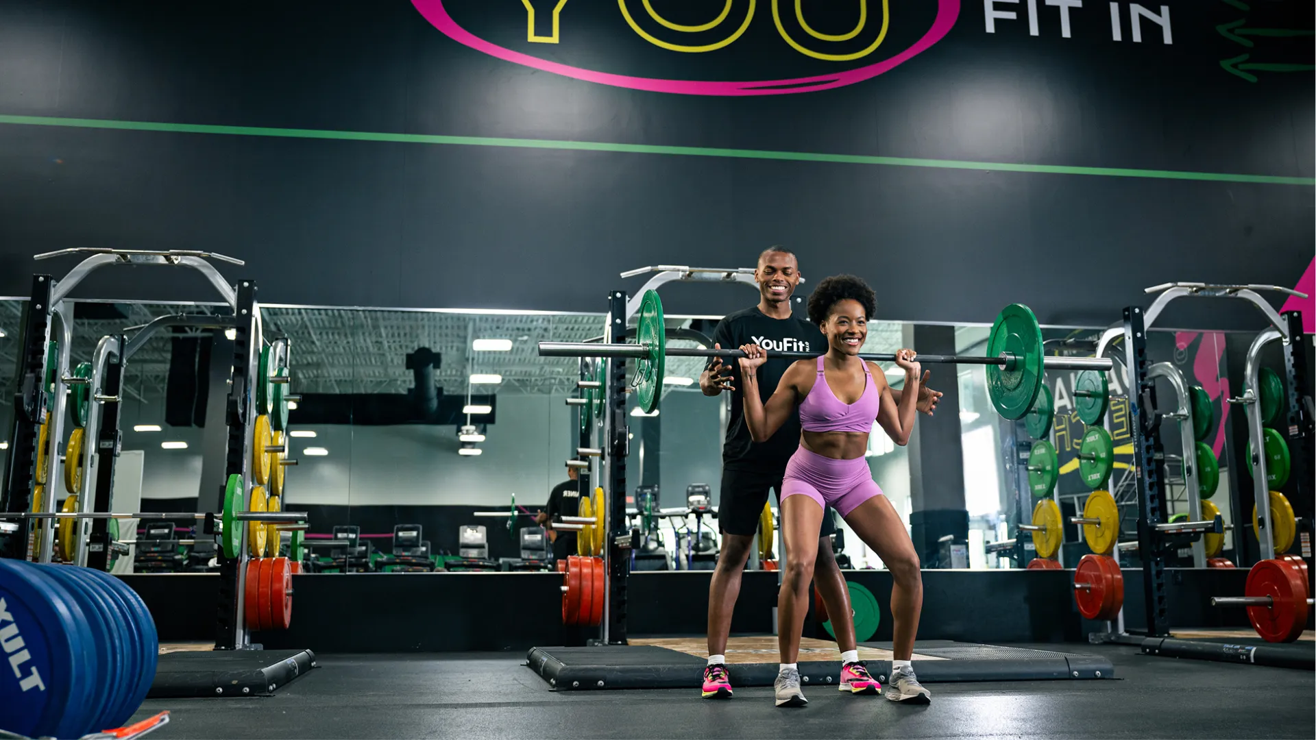 Young woman lifting barbell with trainer support in a spacious gym with motivational wall text