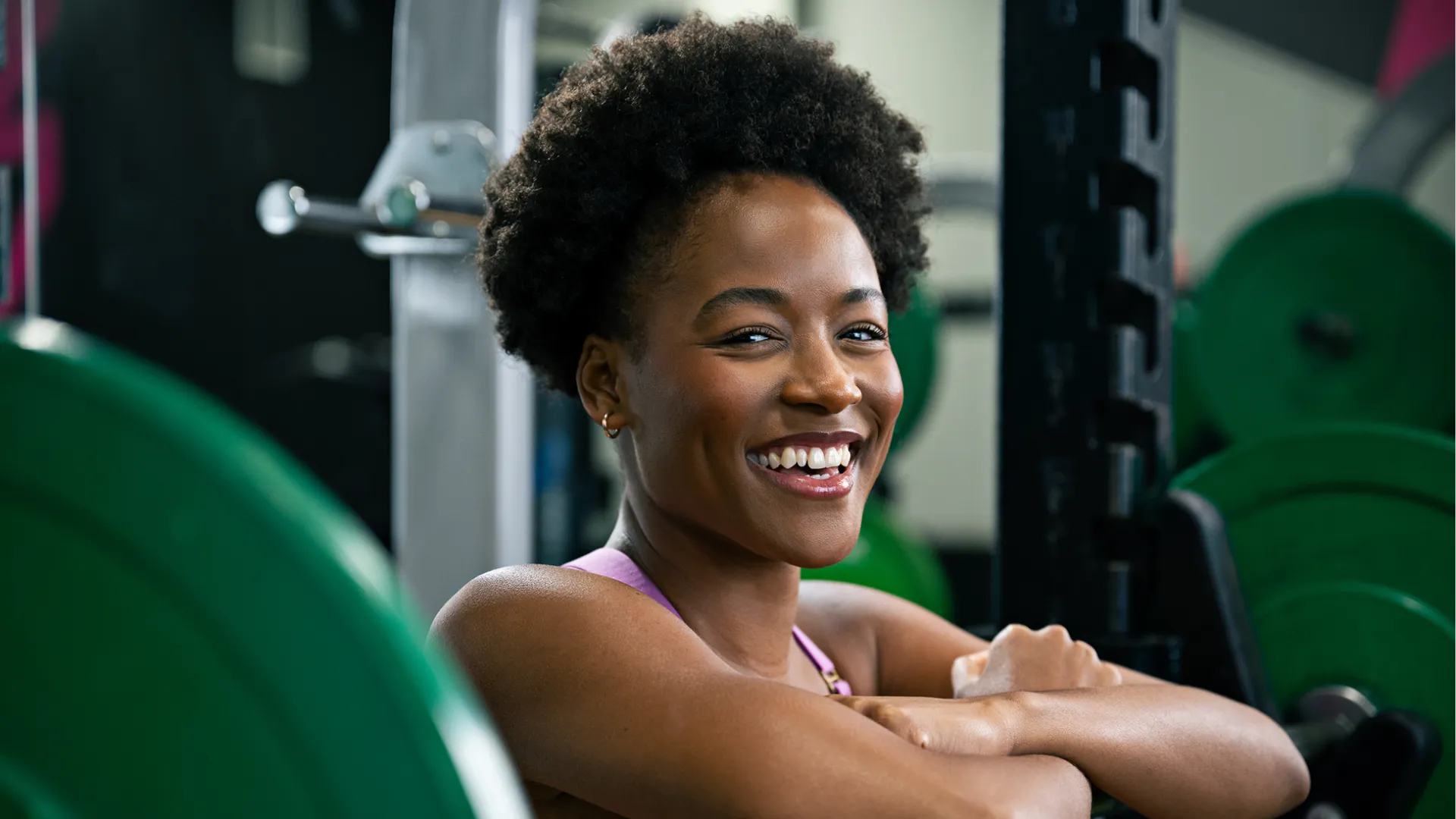 Smiling woman resting on a barbell rack in a gym surrounded by weight plates and fitness equipment.
