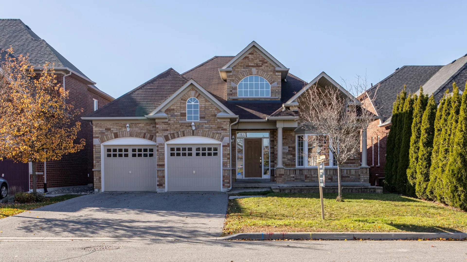 Suburban two-story brick house with double garage doors, front lawn, bare trees, and clear sky.