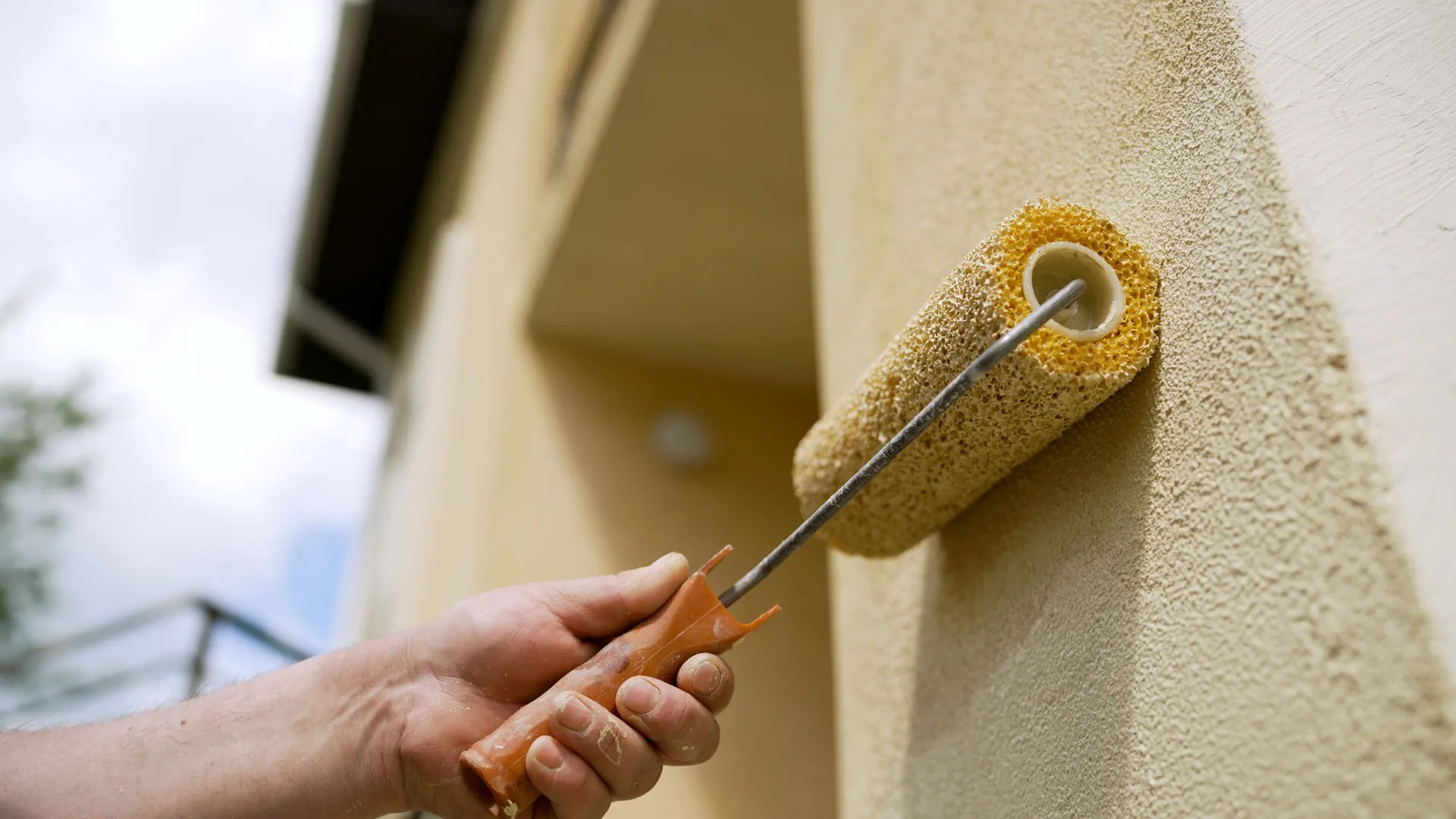 Hand applying beige textured paint on wall with roller brush during exterior home painting.