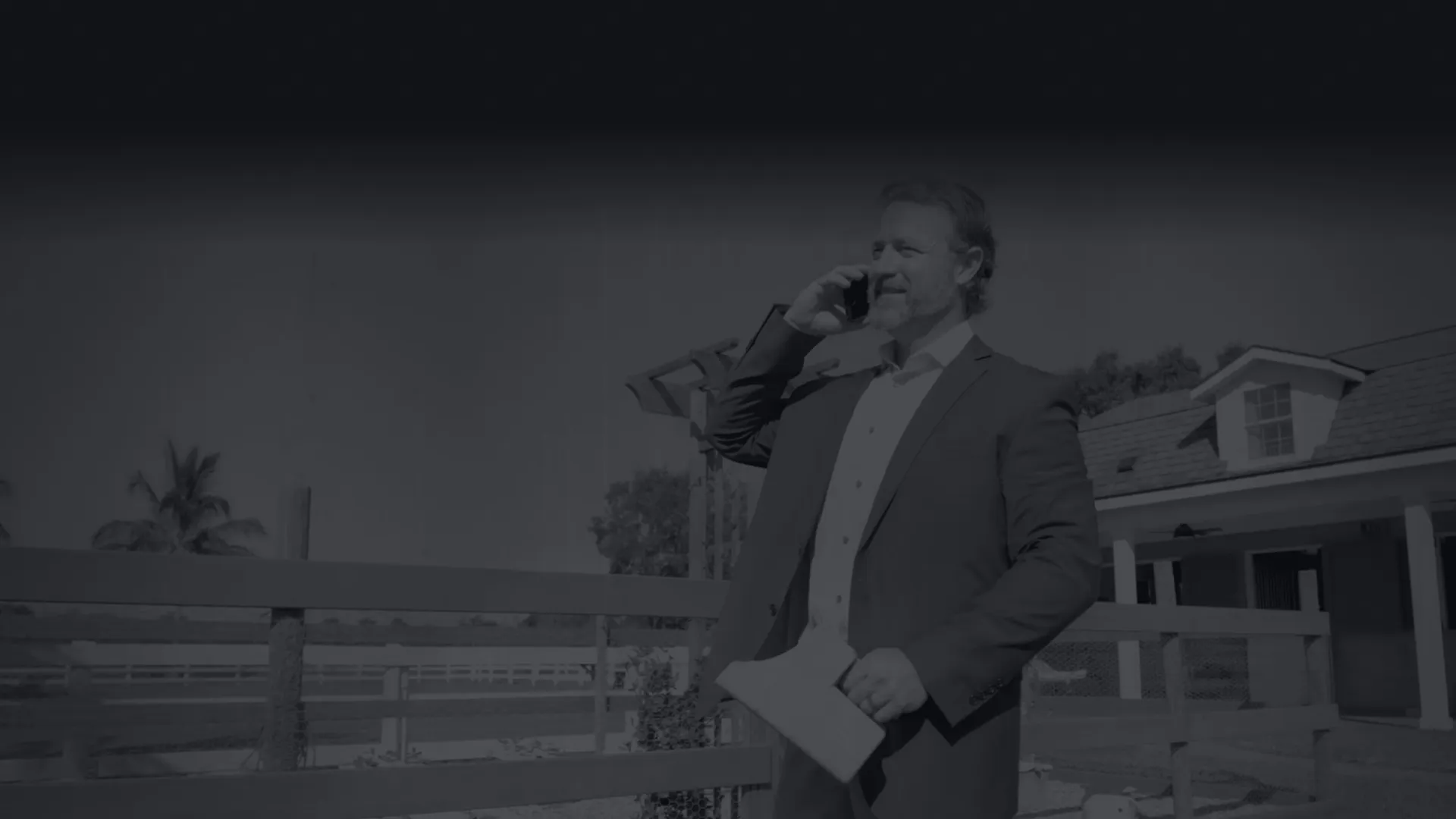 Real estate agent talking on phone holding papers standing outdoors near a house and wooden fence.