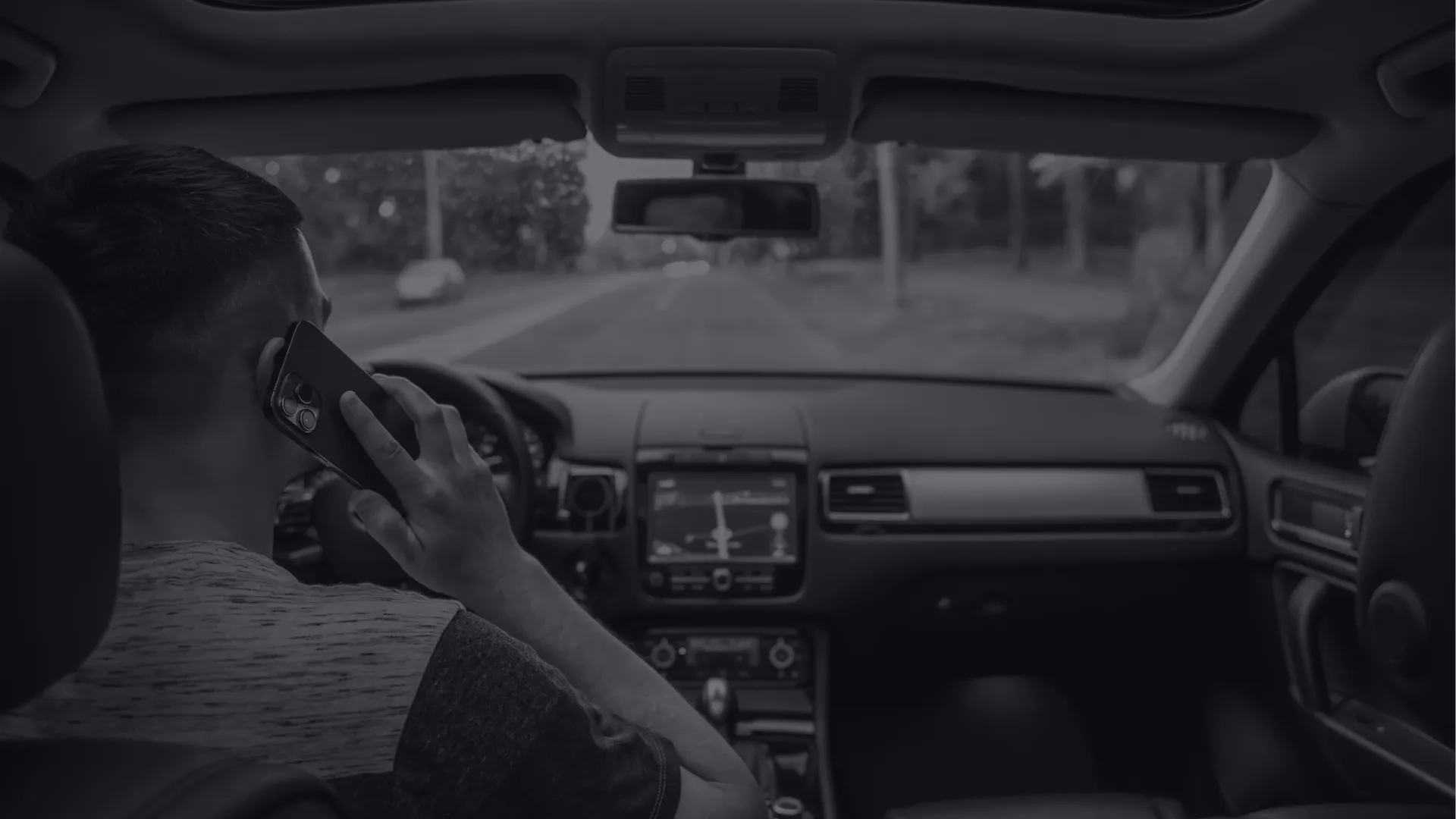 Man inside car talking on phone while driving on an empty road in daylight.