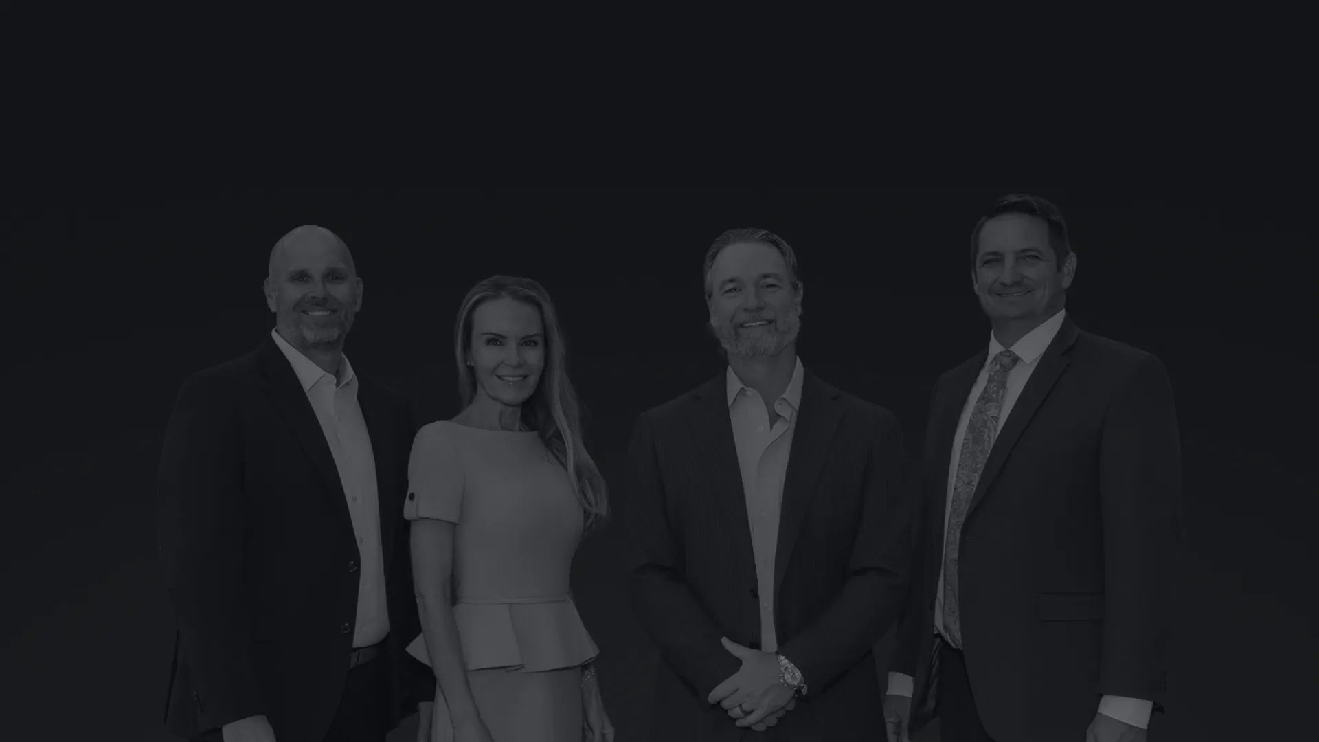 Four professionally dressed businesspeople standing side by side against a plain background, smiling.