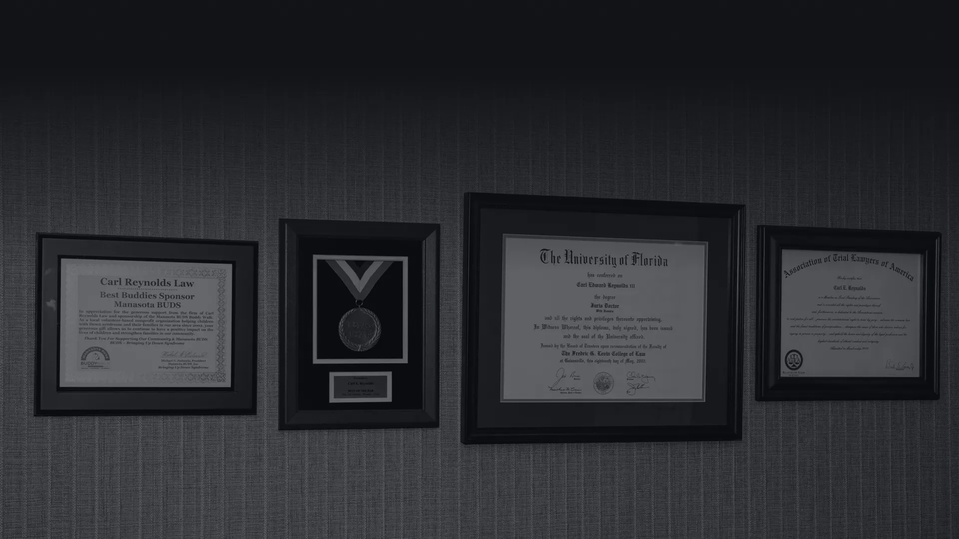Wall displaying framed diploma, certificates, and a medal in black frames on textured gray wallpaper.