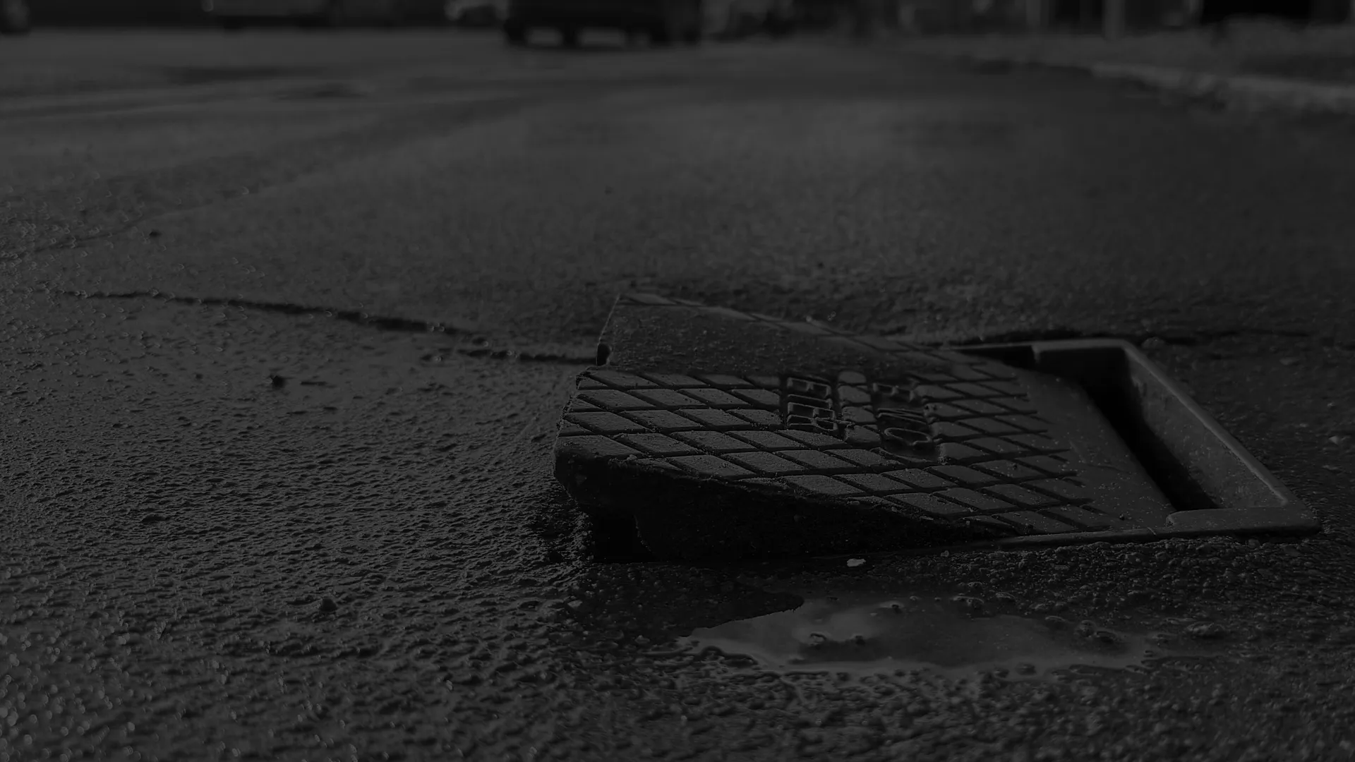 Broken storm drain cover displaced on rough asphalt street with blurred vehicles and buildings in background.