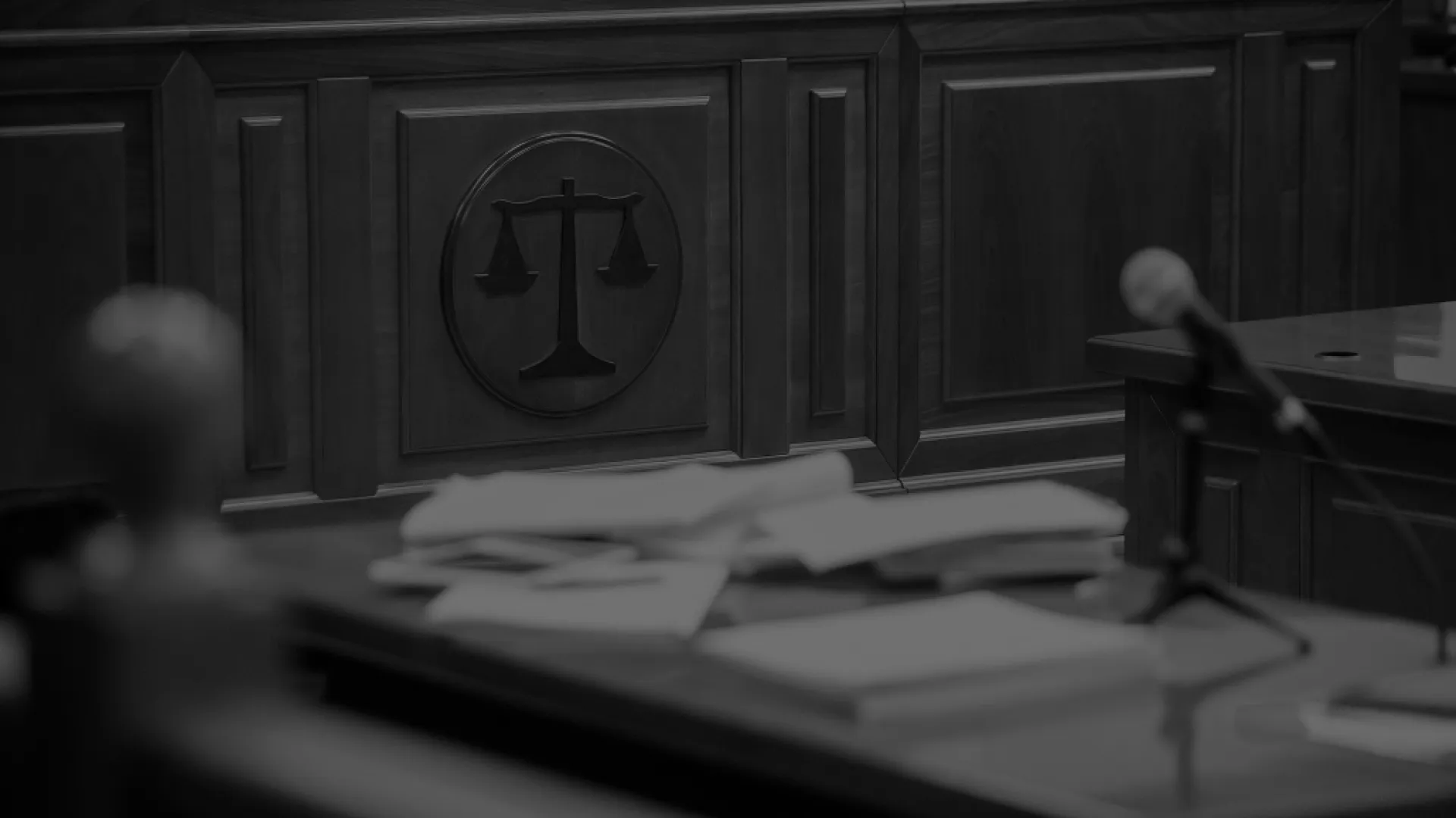 Wooden courtroom interior with scales of justice emblem, papers, and microphone on the table in black and white.