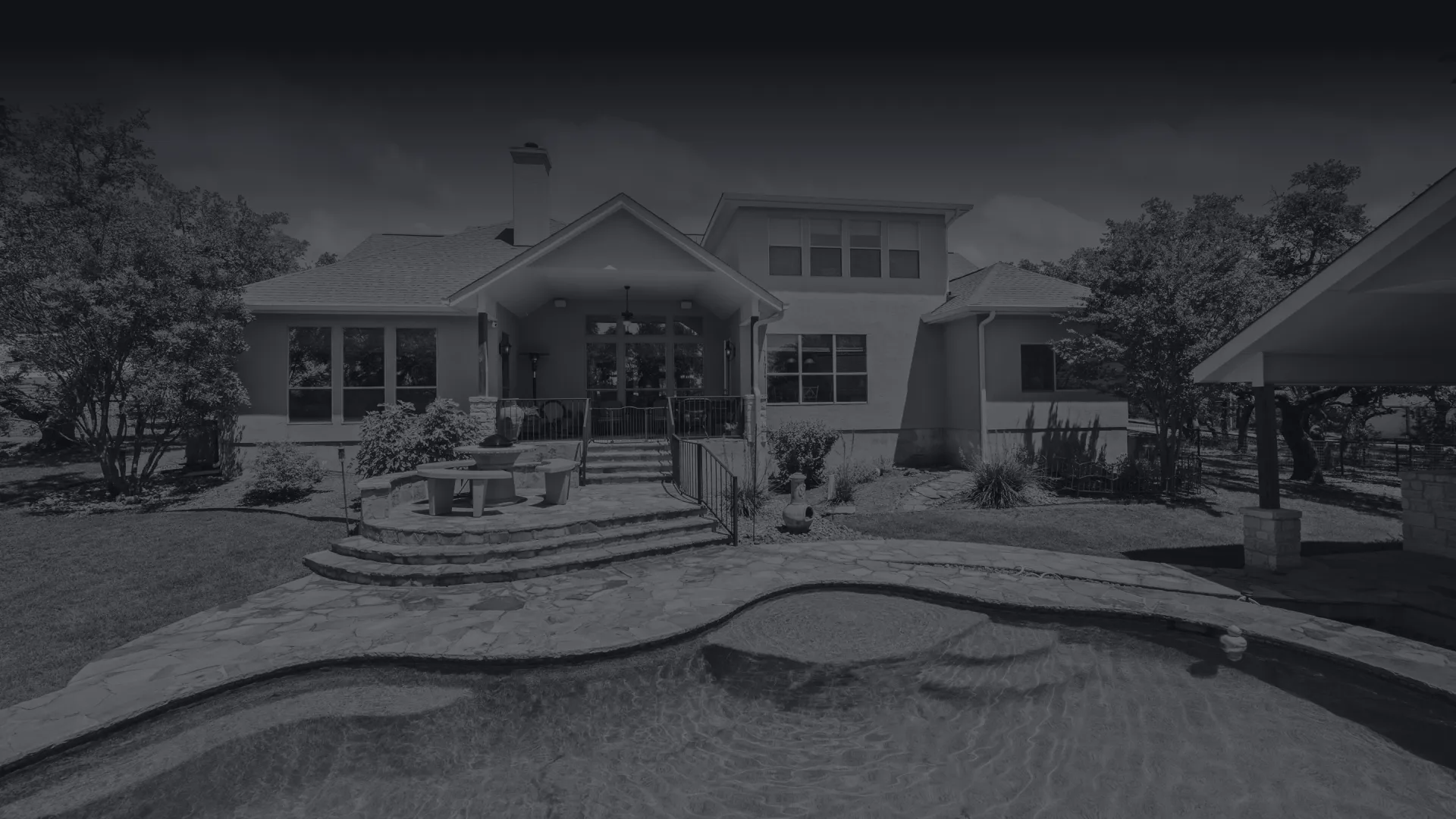 Modern house exterior with a swimming pool, stone patio, outdoor seating, and surrounding trees in daylight.