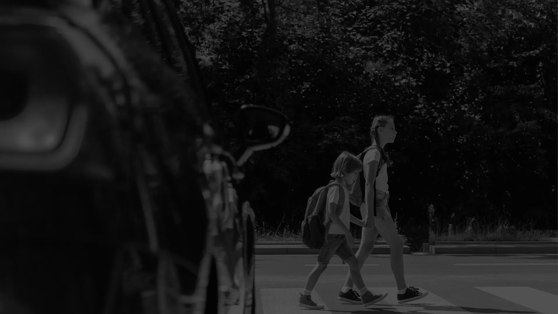 Two children with backpacks crossing a street at a pedestrian crosswalk beside a parked car on a sunny day.