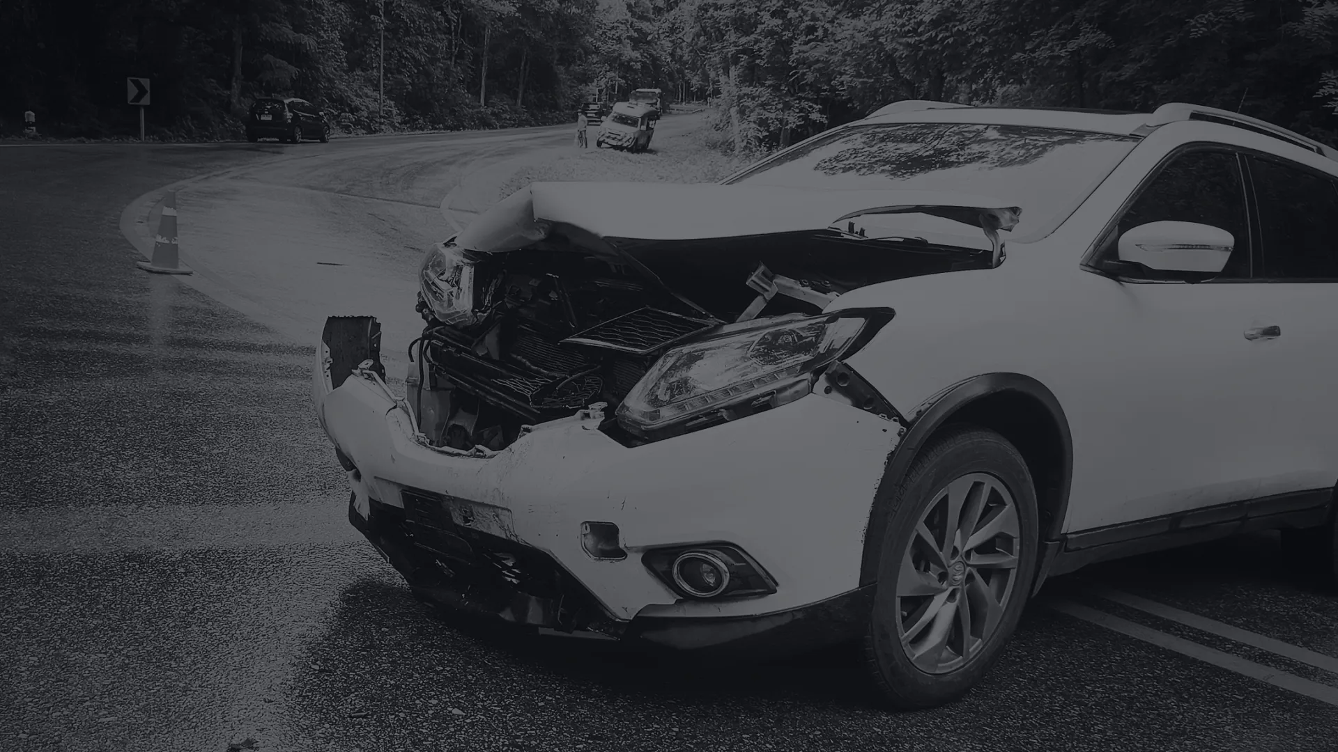 White SUV with severe front-end damage after a crash on a curved, wet road near a forested area.