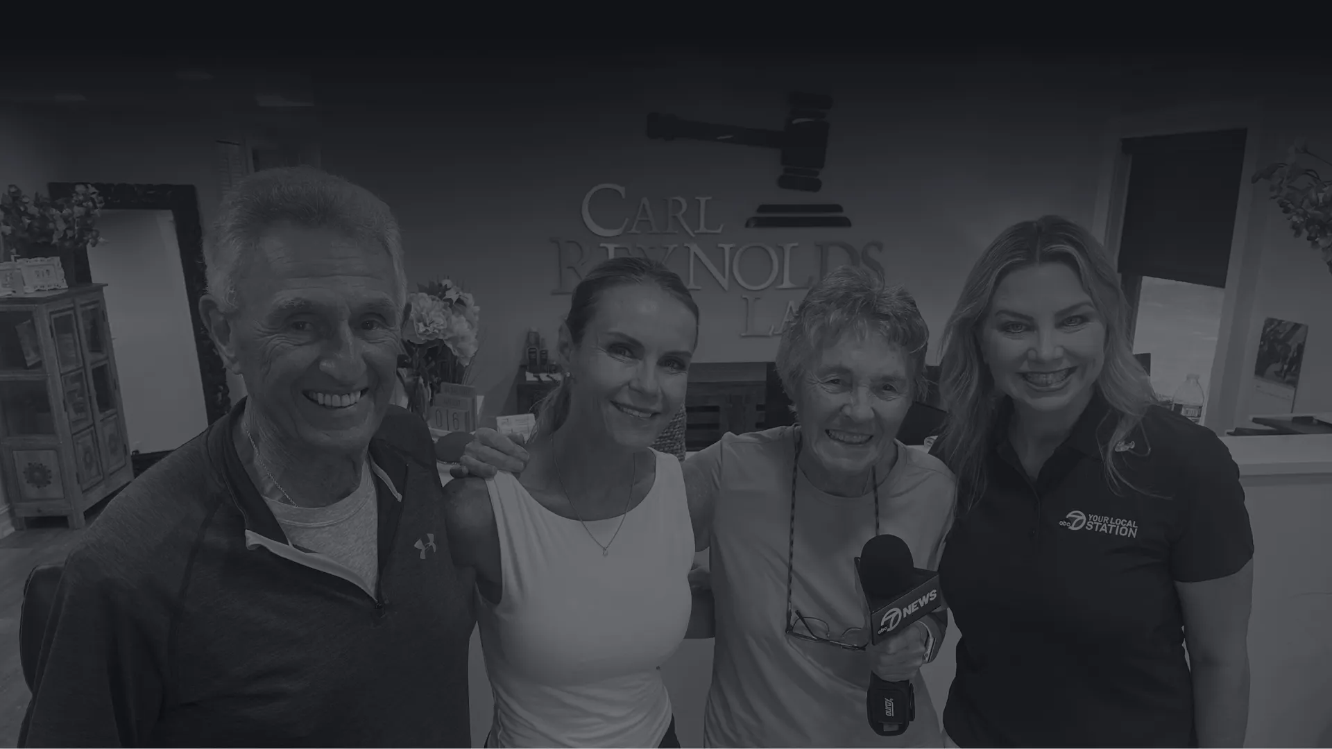 Four smiling people posing together in an office with Carl Reynolds Law sign and a microphone with 7 News logo.