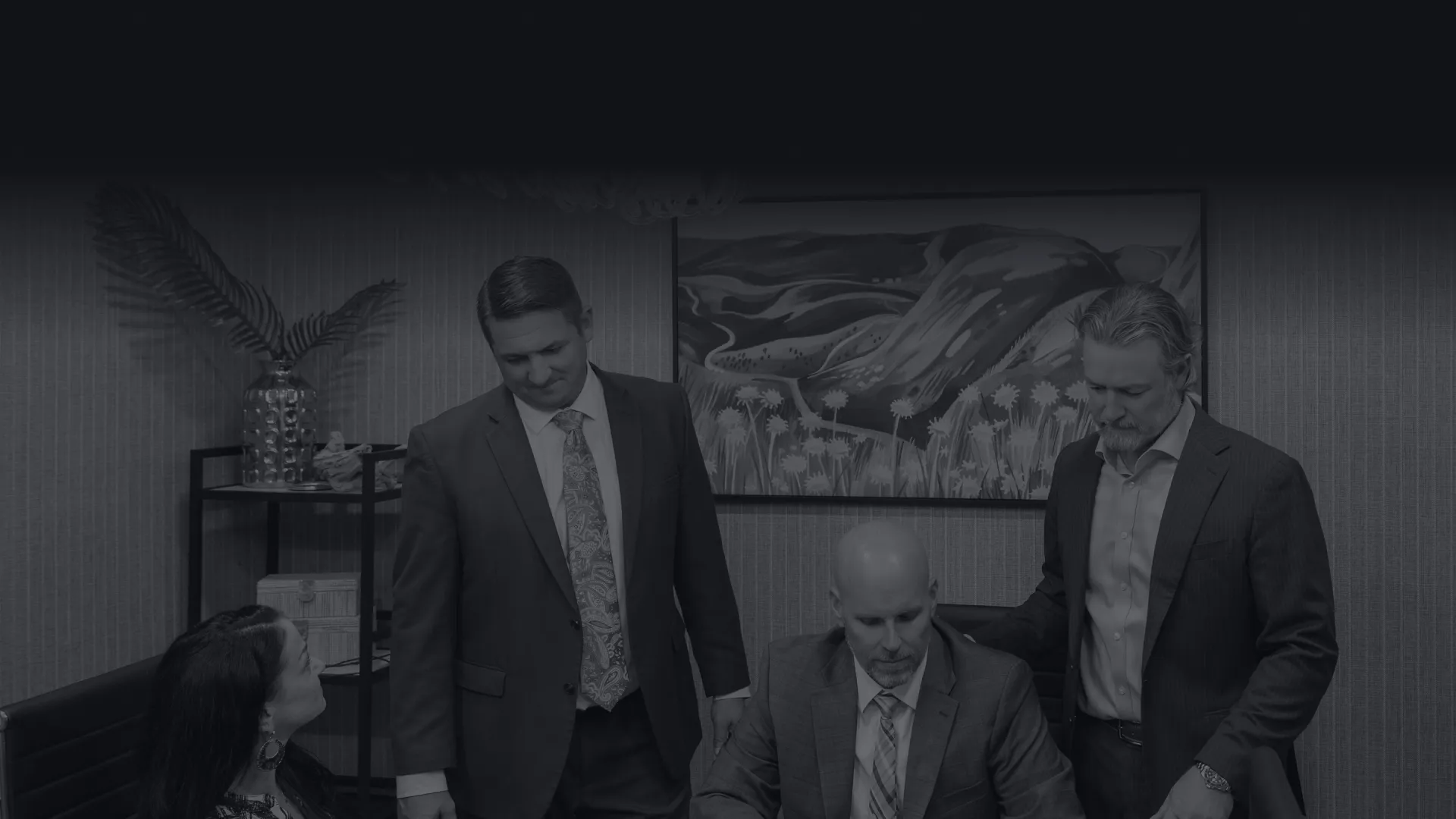 Four business professionals in suits collaborating over a tablet in a modern office conference room.
