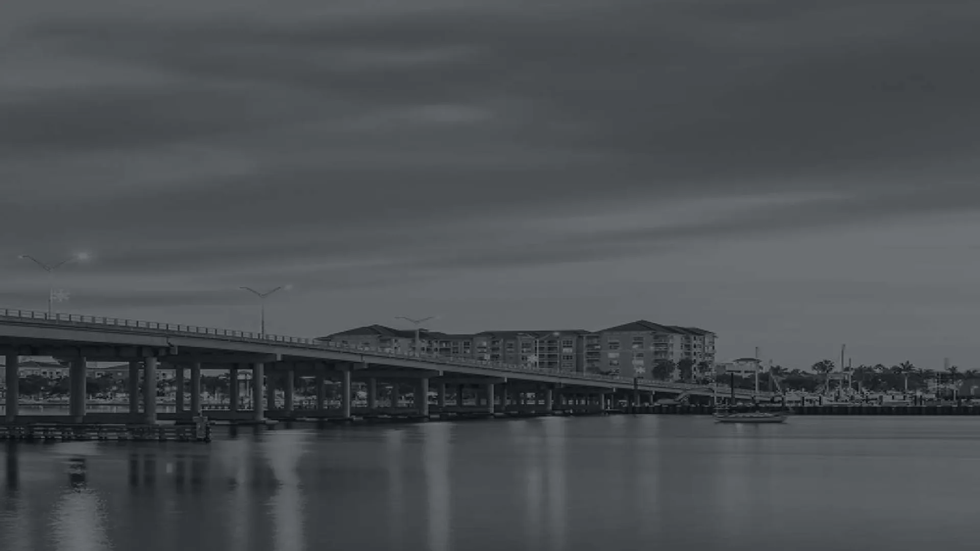 Calm waterfront scene with long bridge extending over water towards distant buildings under cloudy sky.