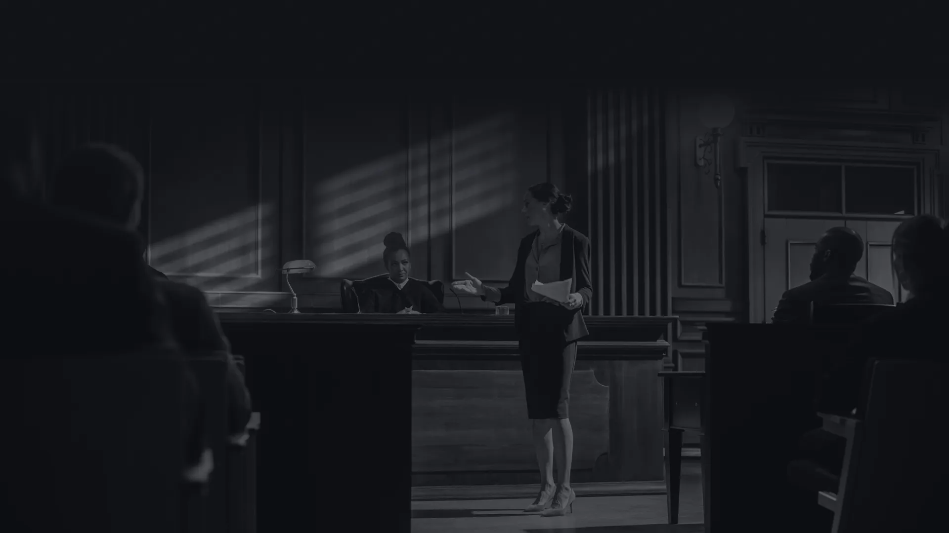 Lawyer presenting case in courtroom with judge and seated audience in black and white setting.