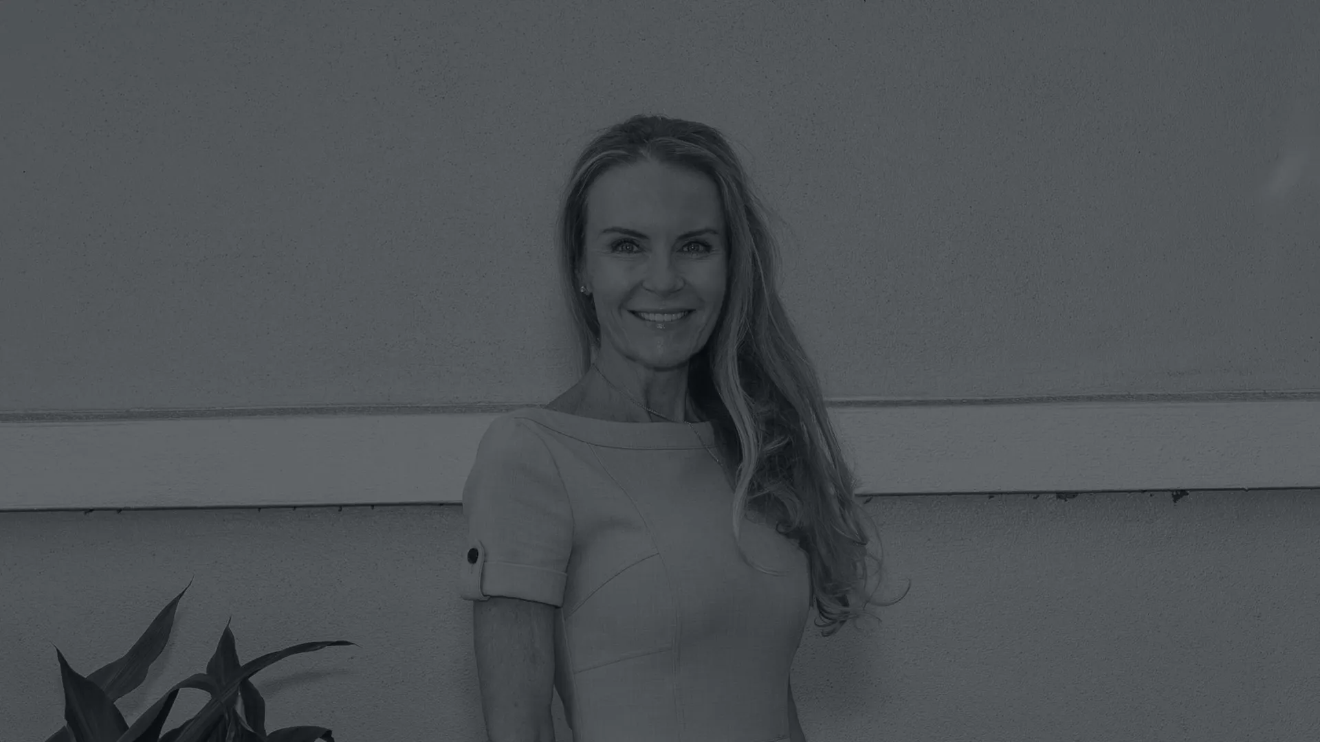 Smiling woman with long hair wearing a fitted dress standing against a wall with plants nearby