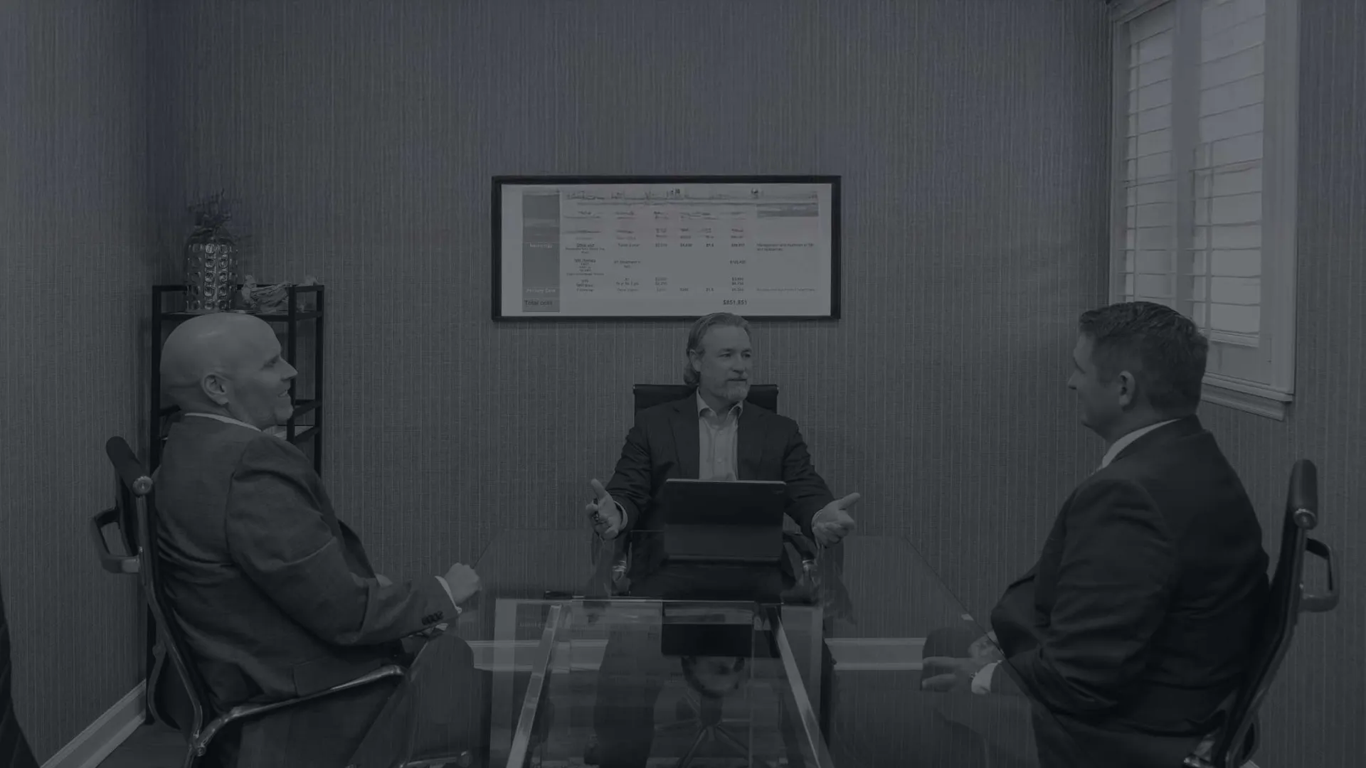 Three businessmen in suits engaged in a discussion around a glass conference table in a modern office.