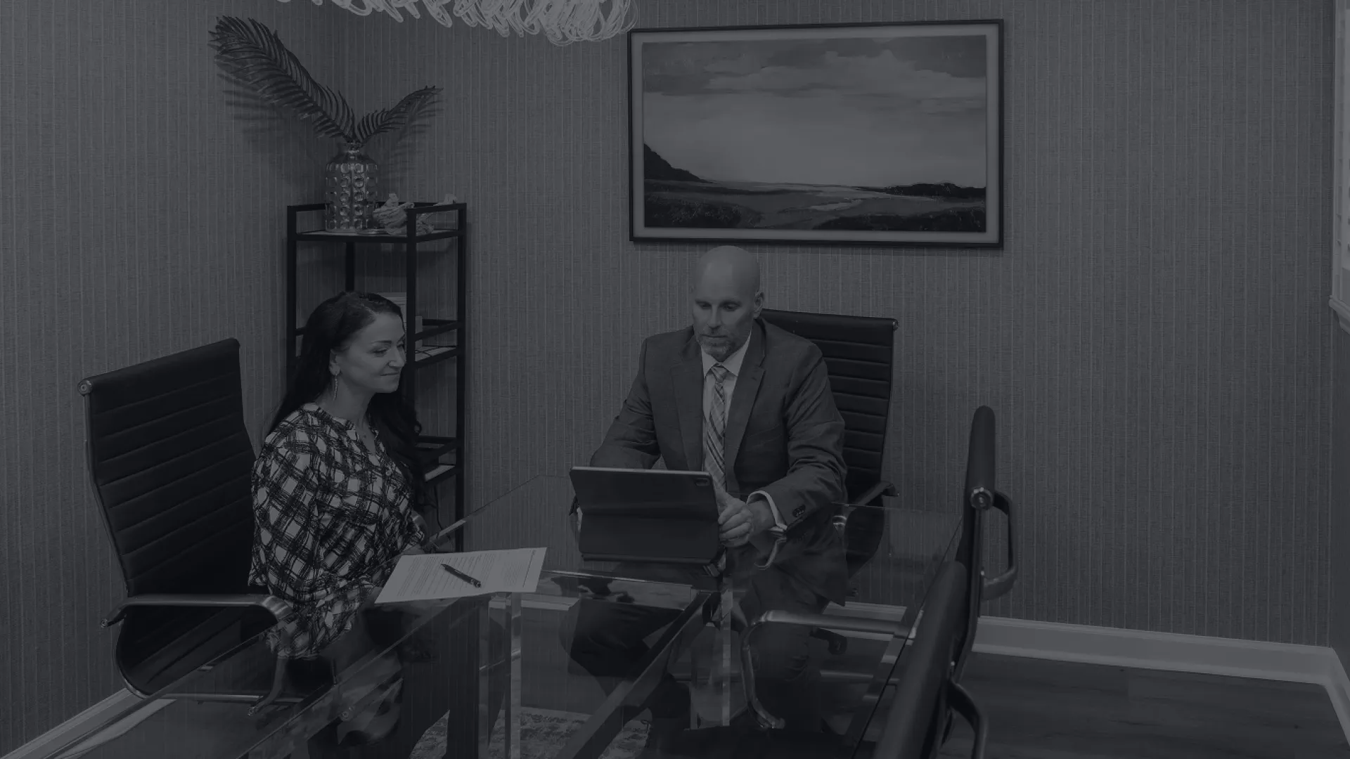 Two professionals in a modern office having a business meeting with laptop and documents on glass table.