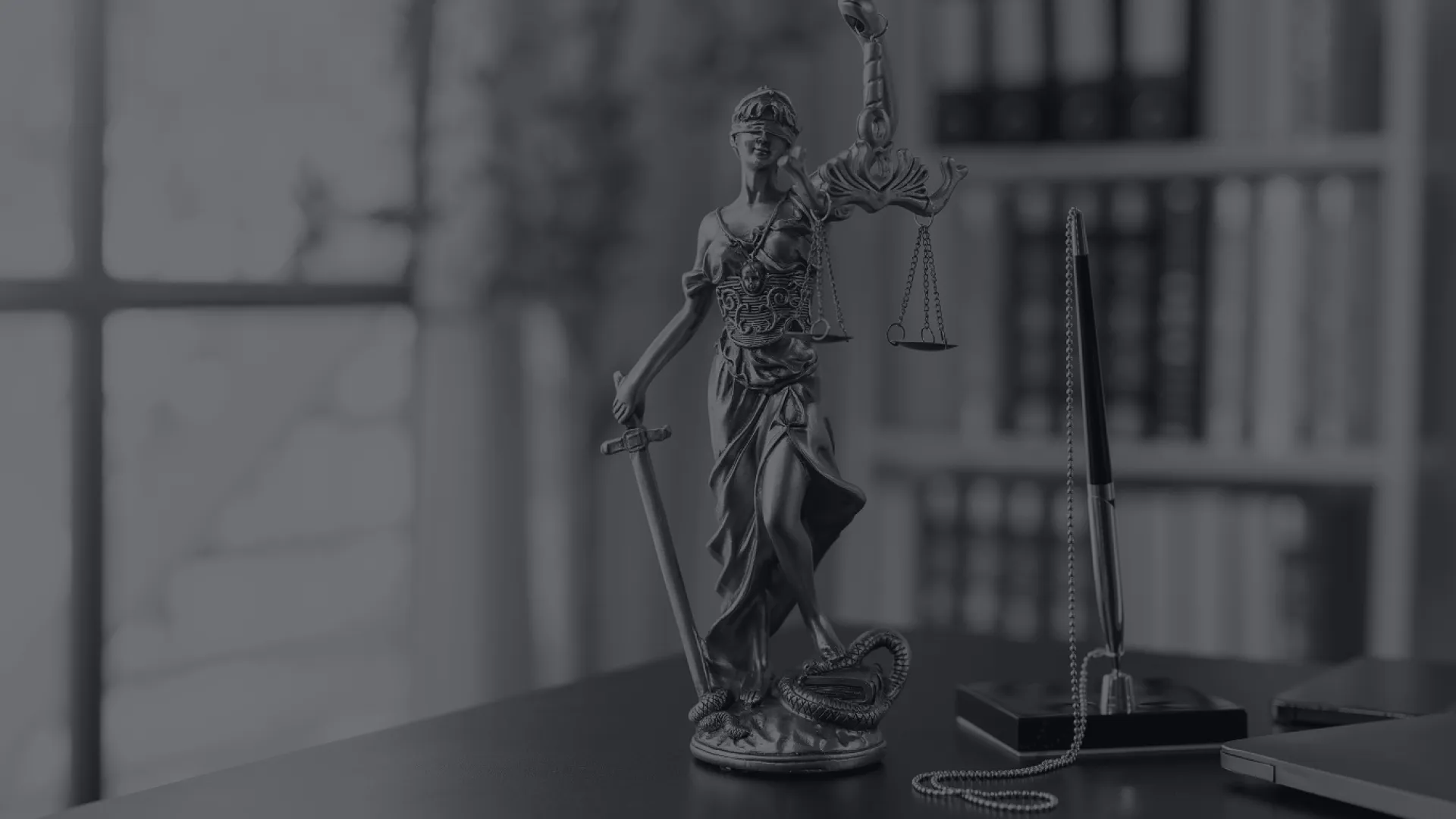 Blindfolded Lady Justice statue holding sword and scales on desk with books and pen in background