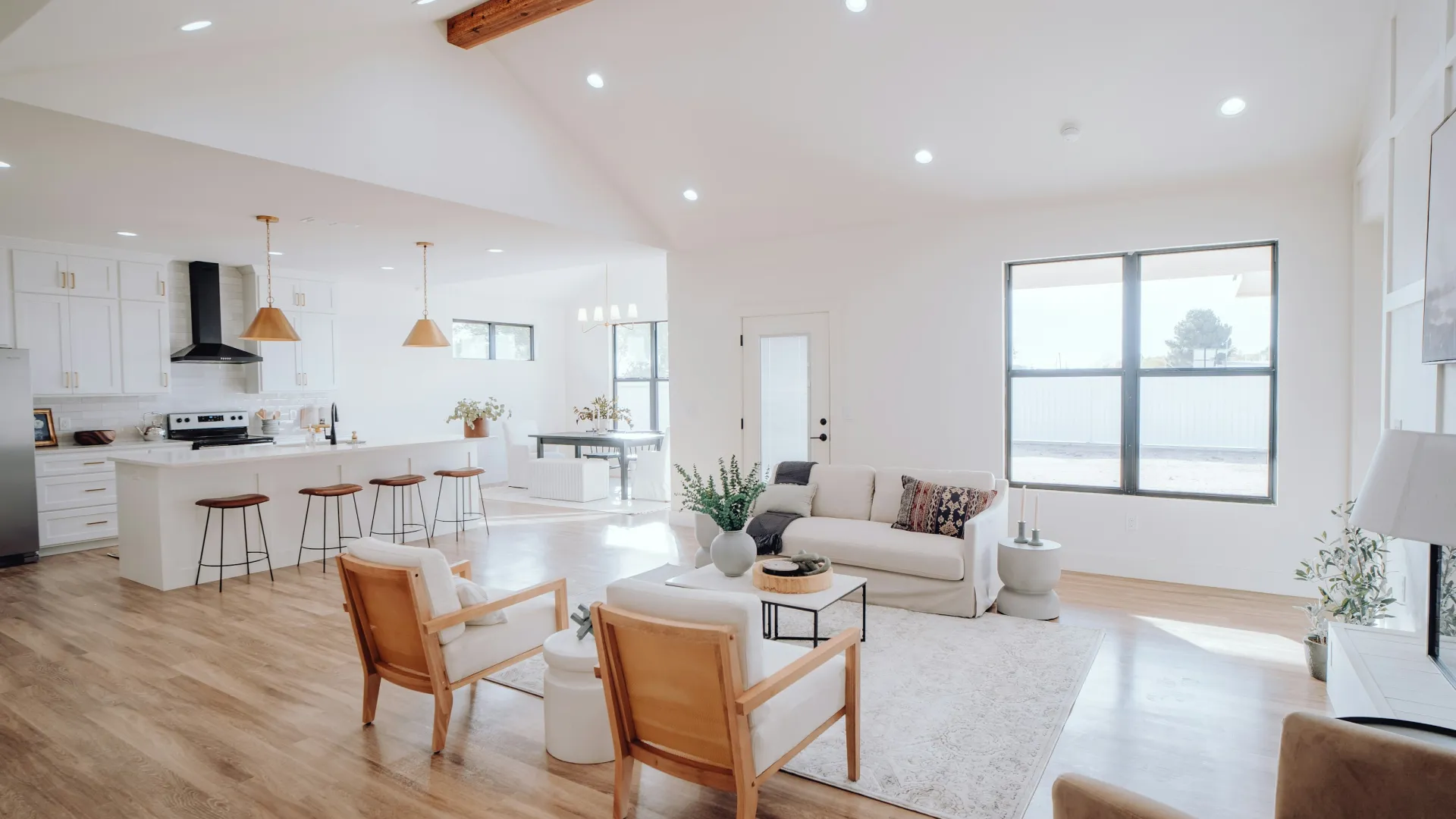 Bright open-concept living room and kitchen with wooden floors, white cabinetry, and modern furniture under exposed wood beams